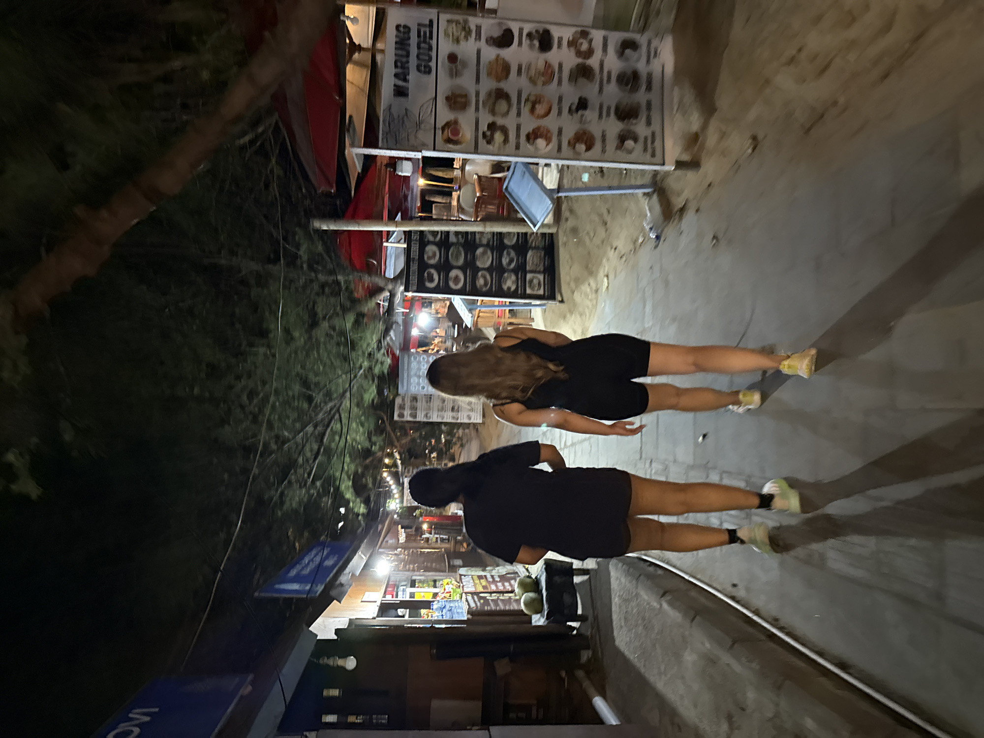 Two women walk away from the camera down a paved path at night. They are walking past shops and stalls on the left, and what appears to be a dining area with umbrellas on the right. The woman on the left wears a black t-shirt and black shorts with light-colored sneakers. The woman on the right wears a black tank top and black shorts with yellow sneakers. The path is illuminated by overhead string lights and lights from the shops. The shops on the left appear to sell various goods, with one stall featuring coconuts. A menu board is visible on the right, displaying food items. The overall environment suggests a bustling night market or street food area in a tropical location, possibly near a beach given the sandy areas visible. This scene is consistent with the nightlife and street vendors often found in popular areas of Denpasar, Indonesia.