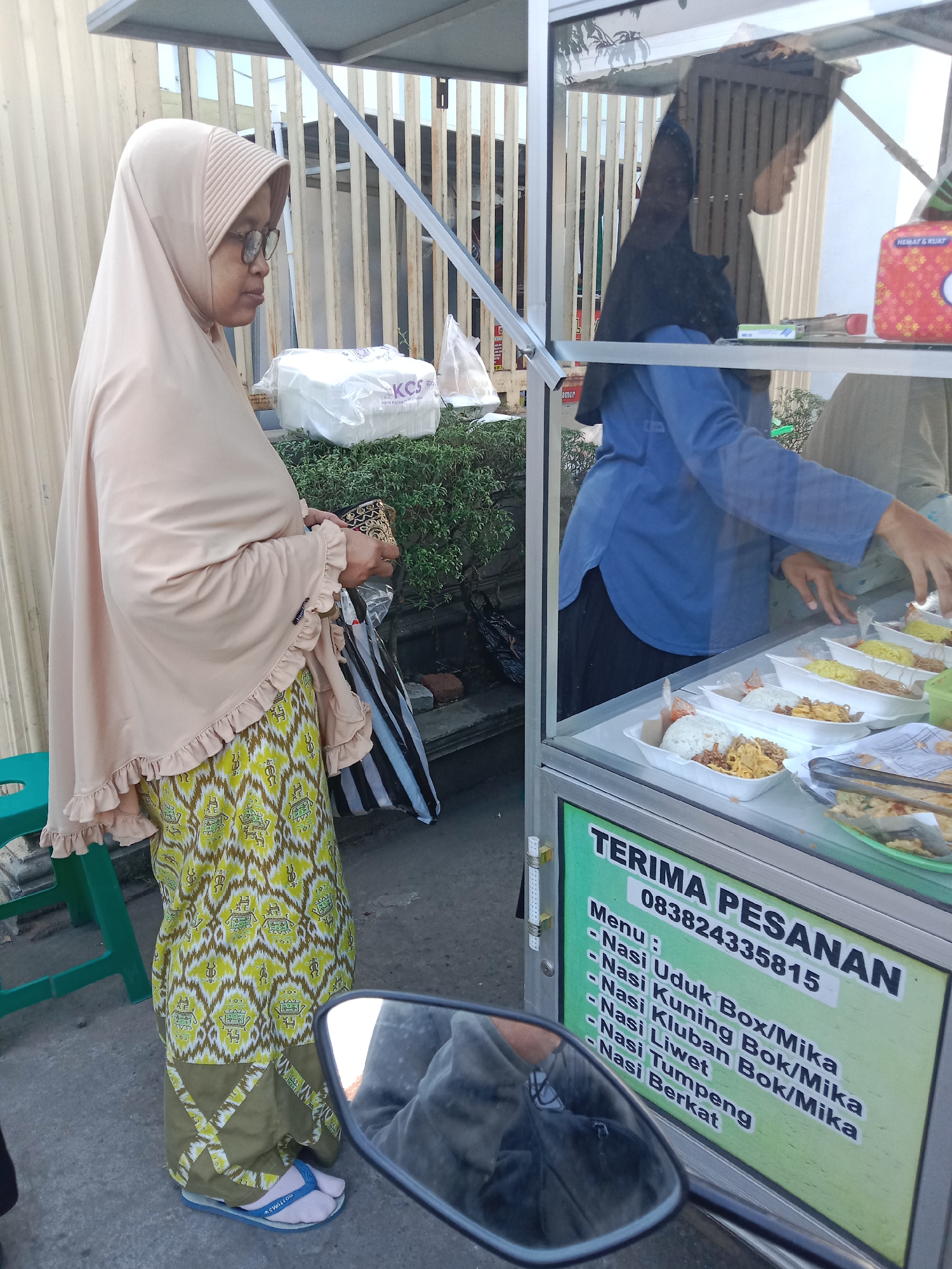 In Pilang, Indonesia, a woman wearing a beige hijab and a patterned skirt stands in front of a food stall. The food stall has a glass display case filled with various boxed meals. Another person, wearing a blue shirt and a black hijab, is seen behind the glass, attending to the food. A menu board is attached to the front of the stall, listing different rice dishes. The scene appears to be an outdoor setting, possibly a street market, with a scooter's mirror visible in the foreground, reflecting a portion of the scene.