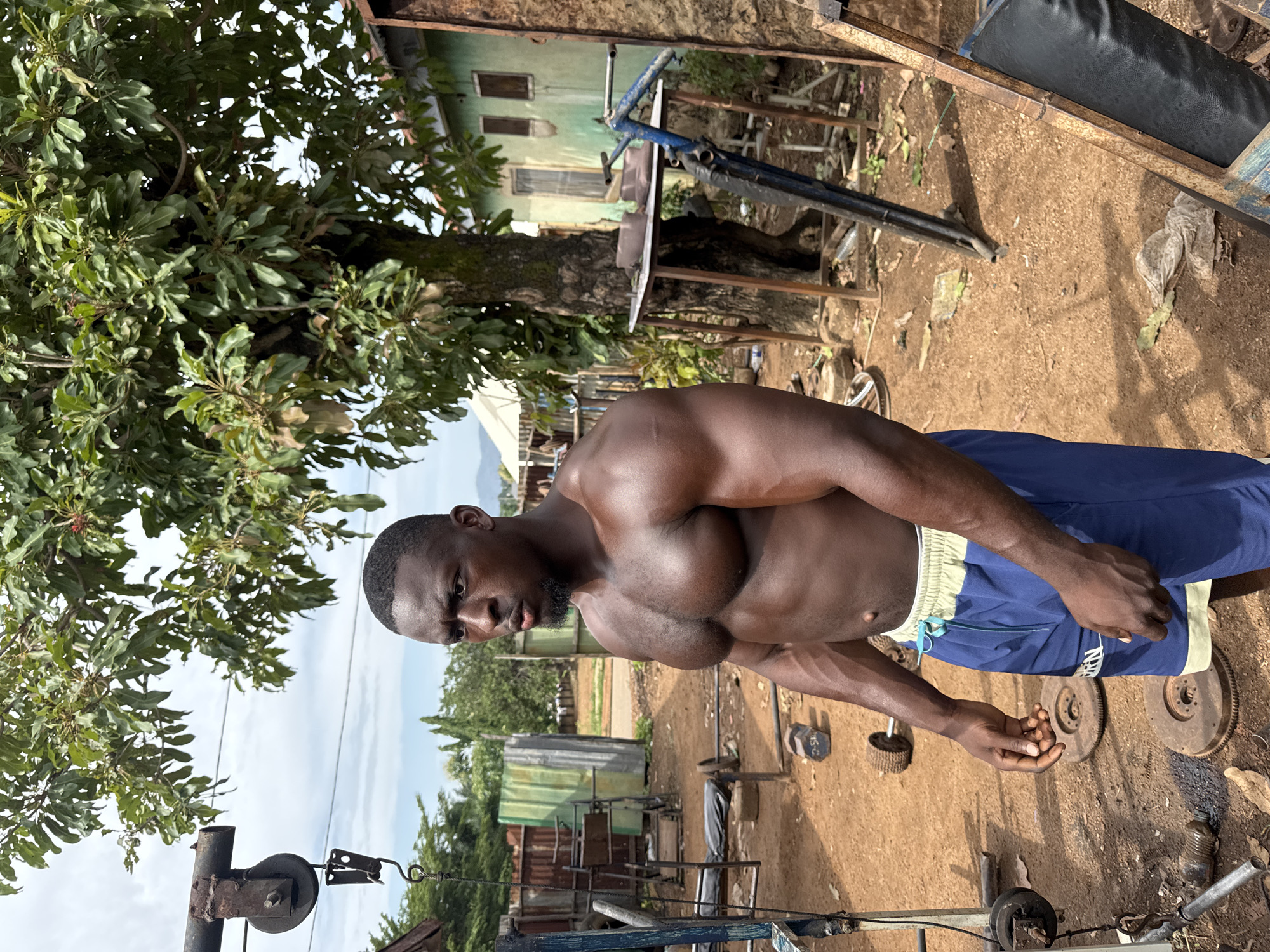 A muscular, shirtless Black man is the focal point of the image, positioned in what appears to be an outdoor gym in Akwanga, Nigeria. He is wearing blue shorts with a light-colored waistband. The gym is makeshift, consisting of simple weightlifting equipment constructed from metal pipes, pulleys, and what appear to be car parts used as weights. The ground is dirt. In the background are trees, a partially visible building, and other elements of the environment. The overall setting is an area that seems to be a low-income or less developed part of Akwanga.