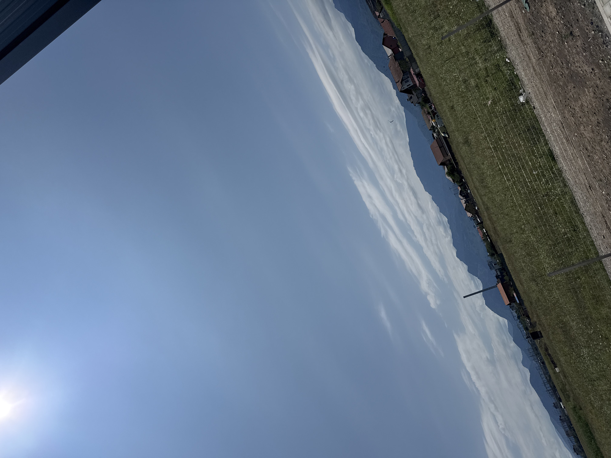The image depicts a landscape in the city of Sanpetru, Romania. The sky is a clear blue, with some wispy clouds visible. The sun is shining brightly, casting a glare in the upper left corner of the image. In the foreground, a grassy field is bordered by a dirt path and a wire fence. The field appears to be undeveloped land, with some scattered debris. In the middle ground, a cluster of houses with red tile roofs is visible, along with a tall, slender industrial chimney. Beyond the houses, a range of mountains forms the backdrop. The overall scene suggests a rural or semi-rural setting on a sunny day. There are no people or animals visible in the image.