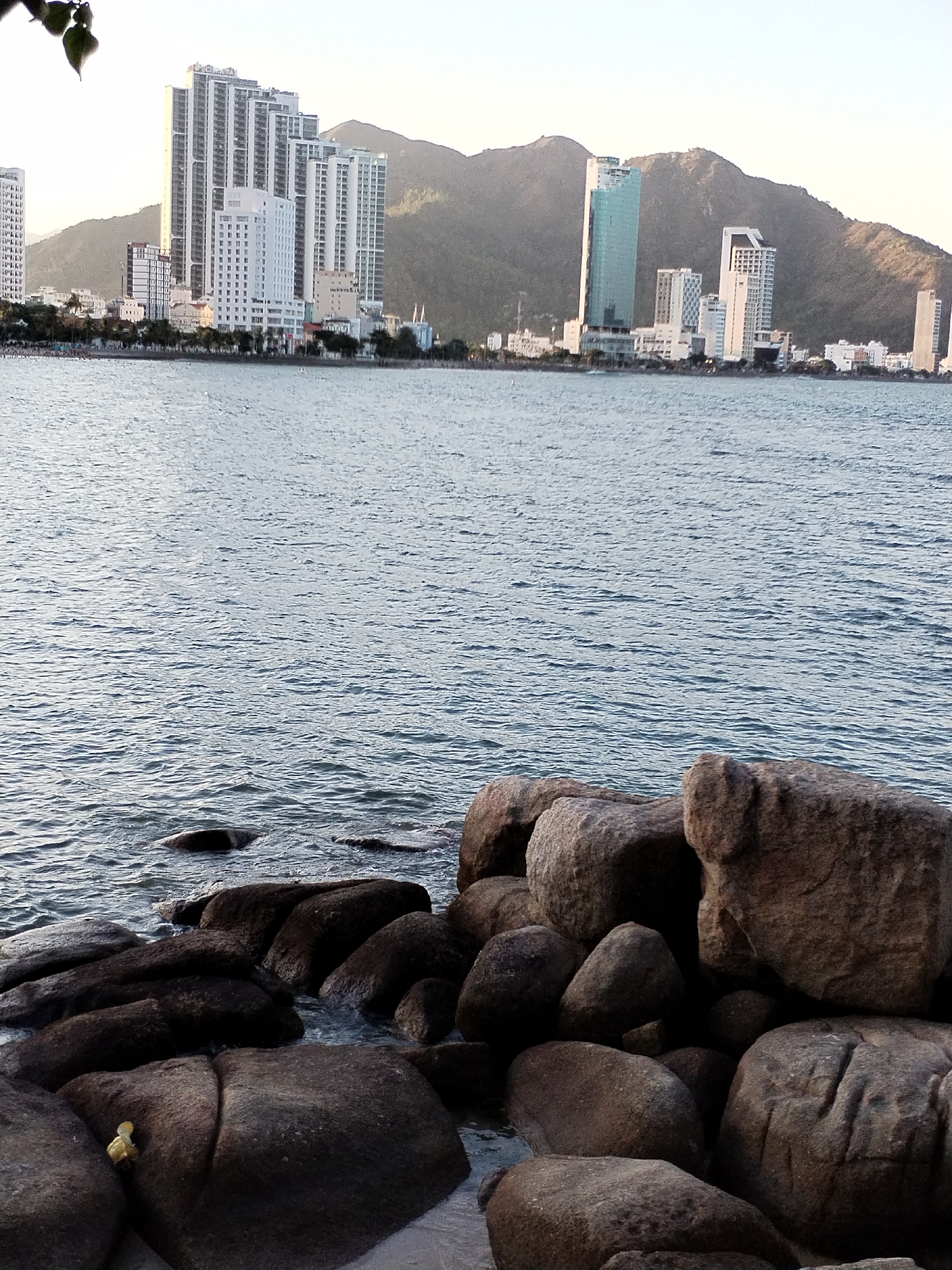 The image shows a coastal cityscape, possibly taken in or near Bac Nha Trang Ward, Vietnam. The foreground features dark-colored rocks, partially submerged in water. A body of water extends toward the horizon, where a skyline of modern buildings is visible. A mountain range rises behind the city. The light suggests it may be early morning or late afternoon.