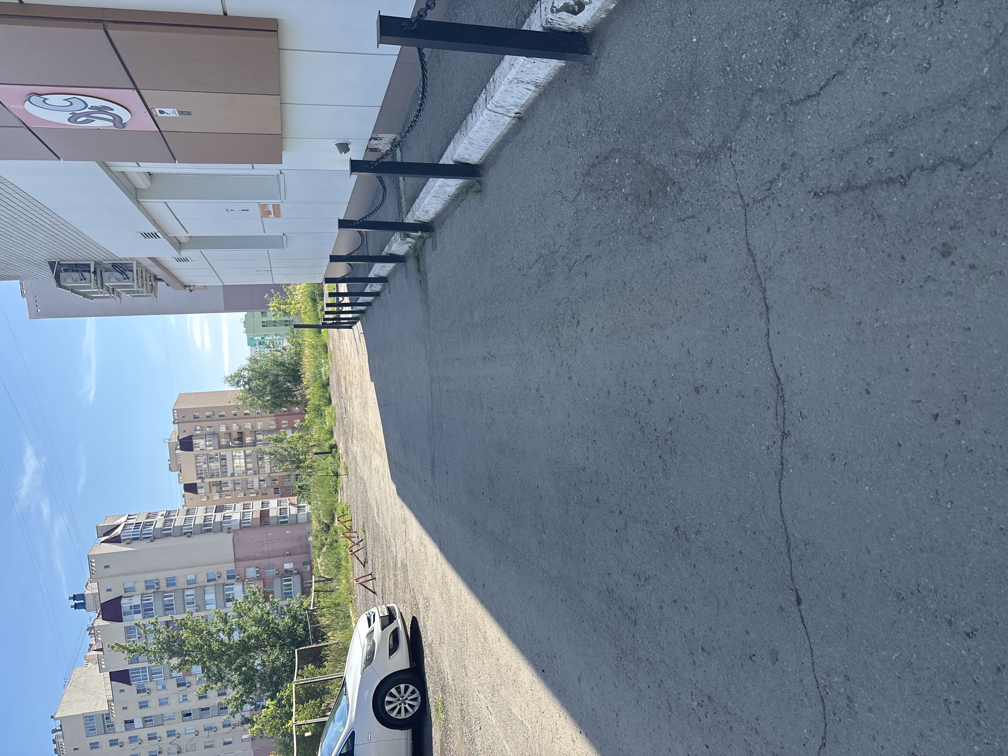 The image shows a parking lot area in front of a modern building in Saratov, Russia. The building has a white and brown facade with a logo on the upper right corner. A black metal chain fence with vertical posts separates the sidewalk from the building. In the foreground, there is an asphalt surface with visible cracks and tire marks. A white car is parked to the left of the frame. In the background, there are multi-story residential buildings and some greenery. The sky is clear and blue, indicating a sunny day.