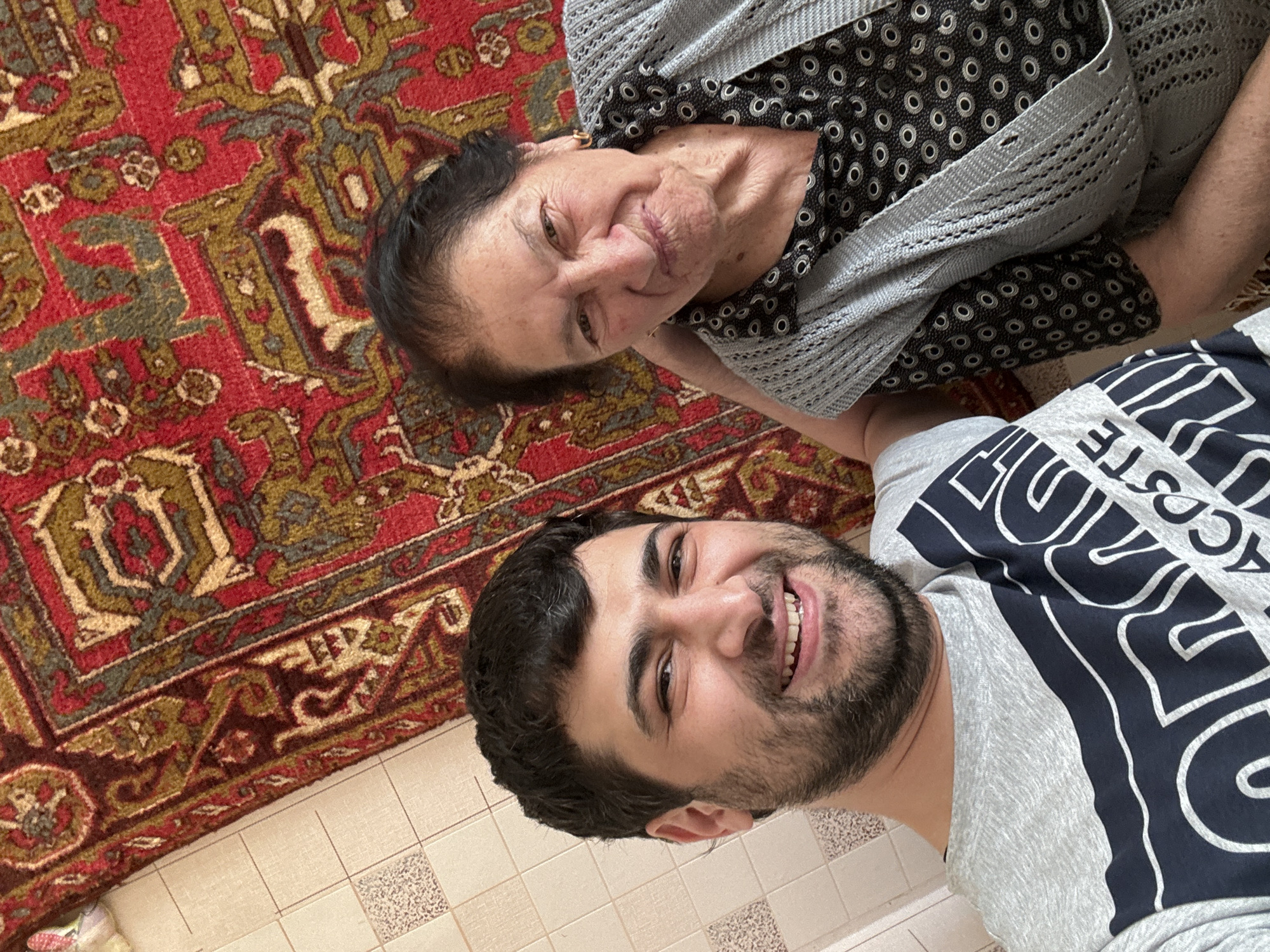 This is a selfie of a young man and an older woman lying on a patterned rug. The woman is on the left, with dark hair and is wearing a black and white patterned top and a grey cardigan. She is smiling and looking at the camera. The man is on the right, with dark hair and a beard, and is wearing a grey t-shirt with dark blue text. He is also smiling and looking at the camera. The rug has a traditional pattern with red, gold, and green colors. The flooring beneath the rug appears to be tiled. The image is taken from a high angle, looking down at the subjects. The lighting suggests it is daytime. The overall mood is cheerful and affectionate. The text on the man's shirt is partially visible, appearing to say "LACOSTE".
