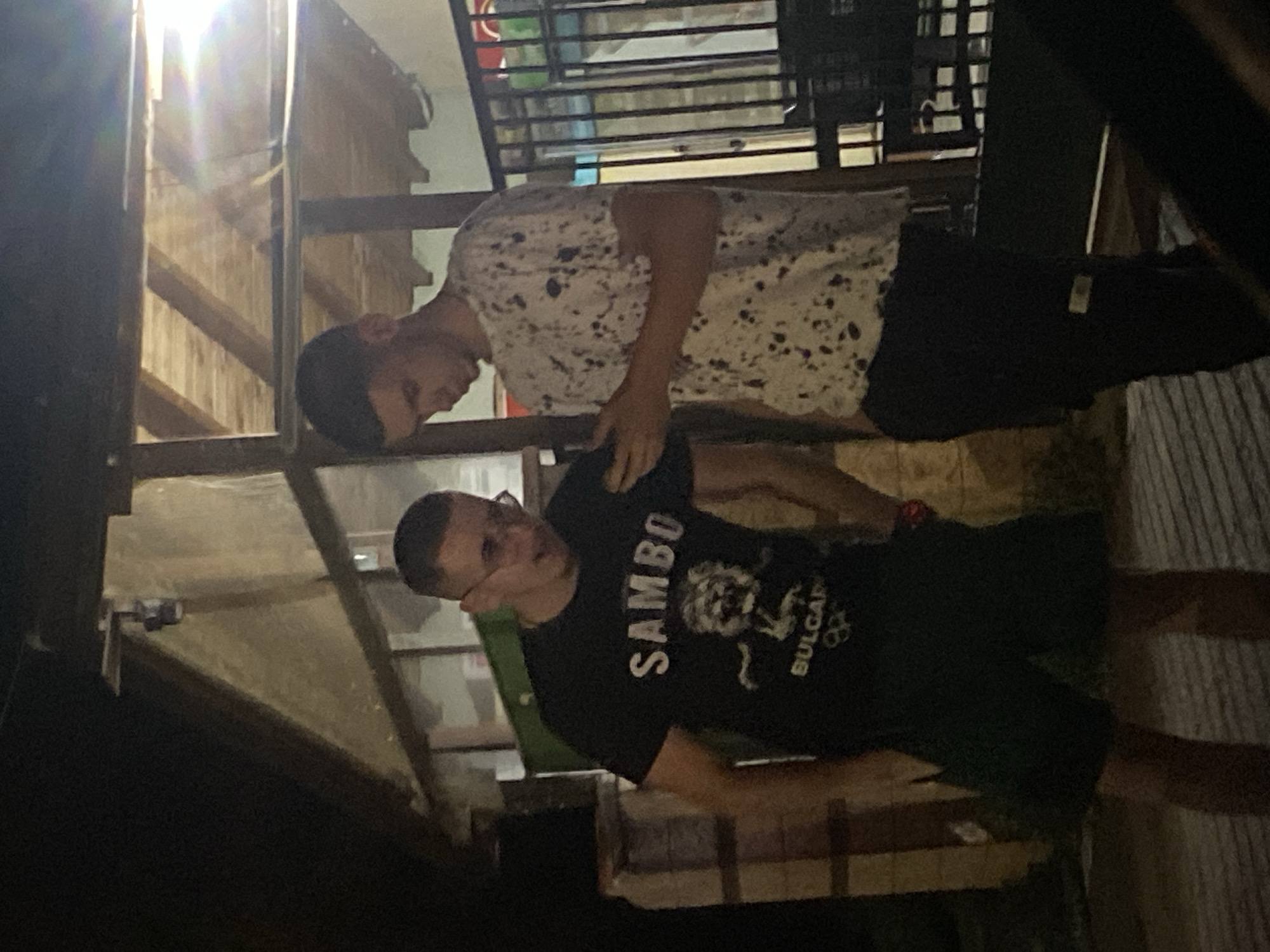 Two young men stand outdoors at night. The man on the left wears a black t-shirt with the word "SAMBO" and a graphic of a lion above "BULGARIA". He also wears dark shorts. He is looking down at a mobile phone he is holding. The man on the right wears a white t-shirt with a black splatter pattern and dark pants. He is also looking down at the phone. They appear to be standing near a building with a wooden overhang. The location is Gorna Malina, Bulgaria.