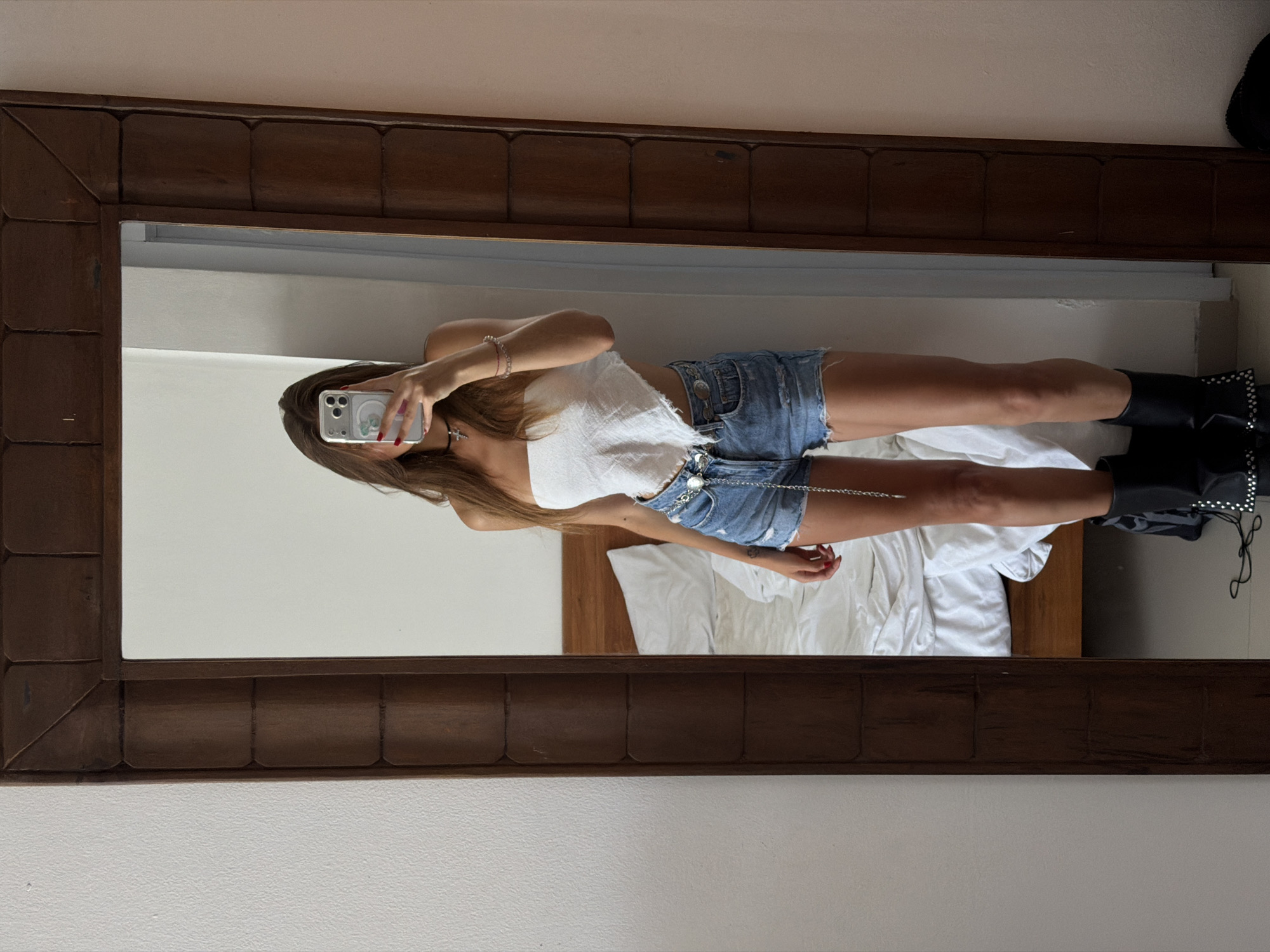 A person is photographed in a mirror within a room. The individual, likely female, is holding a smartphone, presumably taking the picture. The mirror's frame is a dark, textured wood. The person is wearing a white top and denim shorts, accompanied by black boots. A silver chain is draped across the shorts. The background includes a bed with white linens. Given the prompt's mention of North Kuta, Indonesia, it can be inferred the setting is a room within that region.