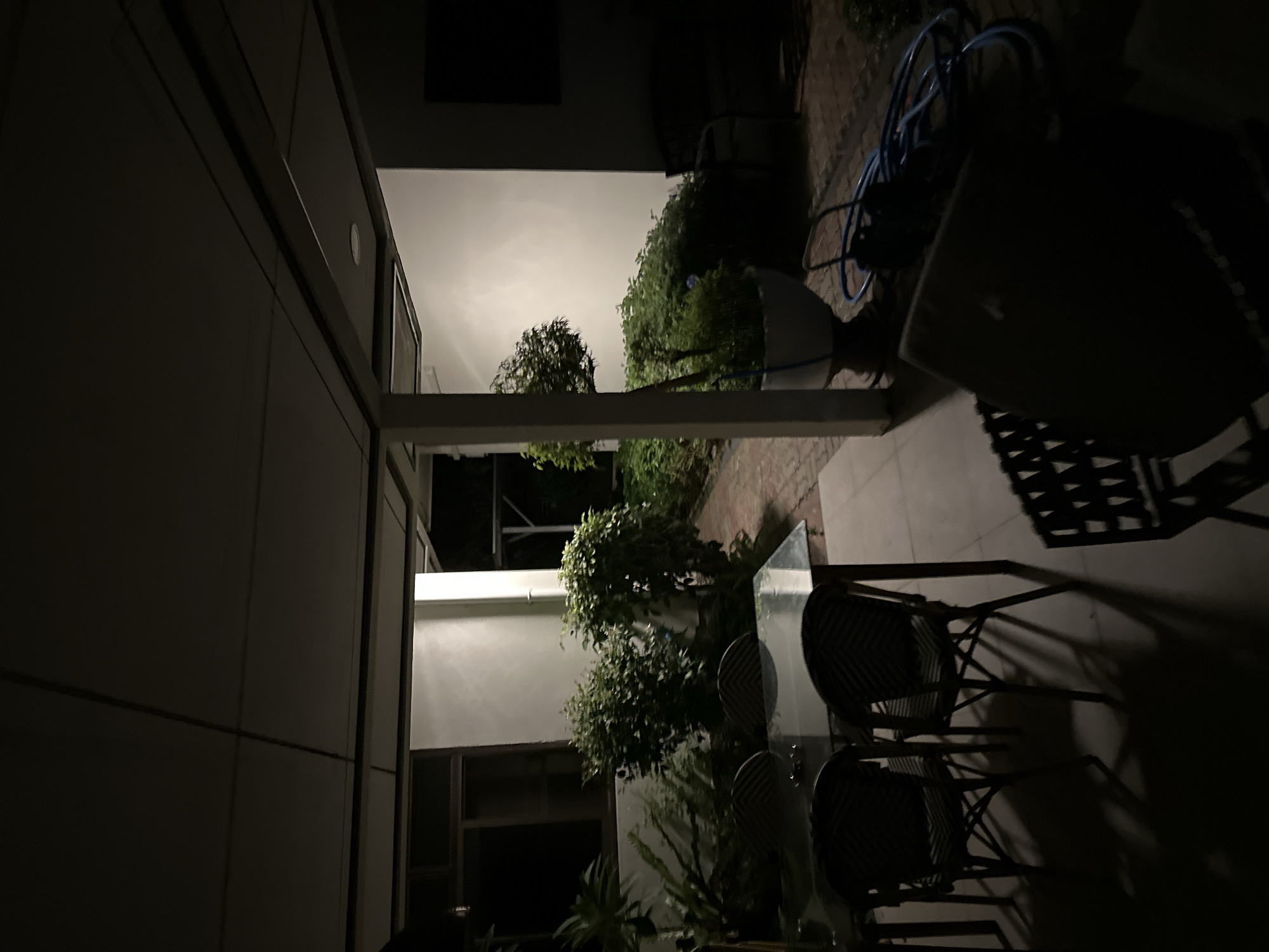 The image depicts an outdoor patio area at night. In the foreground, a glass-topped dining table with four wicker chairs is visible. Beyond the table, lush green plants and bushes are arranged, some in large pots. A blue hose is coiled on the ground to the right, near a large planter. The scene is dimly lit, with shadows cast by the overhead structure and the surrounding foliage. The background shows a light-colored wall and a dark window. No people are present in the image.