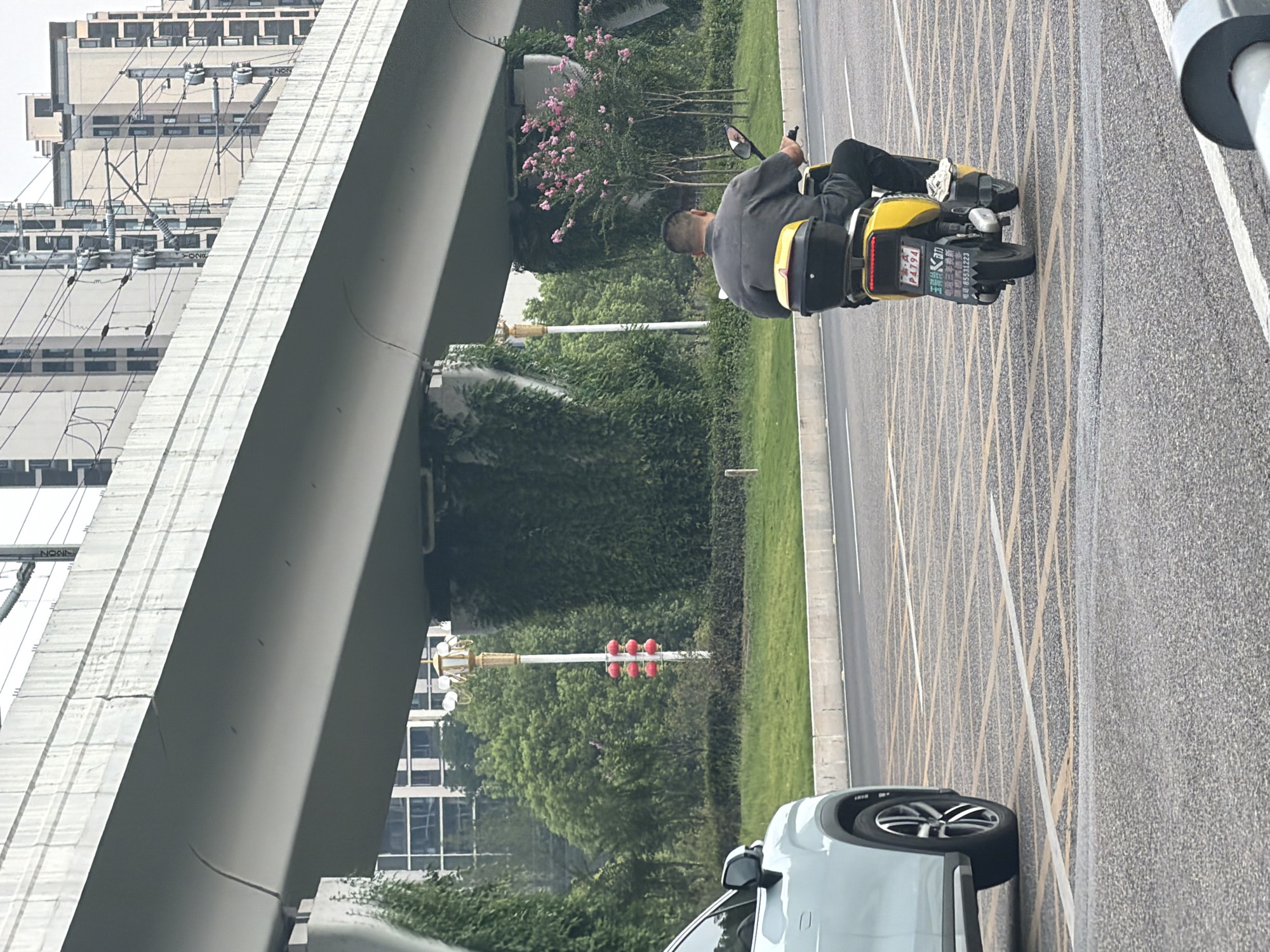 A man is riding a yellow electric scooter in the city of Jiangjin, China. He is wearing a gray short-sleeved shirt and black pants. The scooter has a black storage box on the back. The road is asphalt with yellow and white markings. To the left, a white car is parked. In the background, there is a large overpass supported by pillars covered in greenery. Beyond the overpass, there are modern buildings and power lines. A lamppost with red lanterns is also visible. The weather appears overcast.