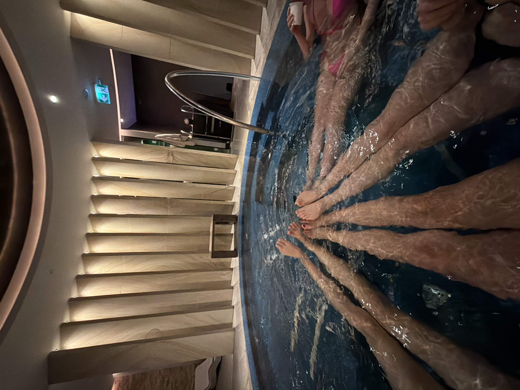 The image shows multiple pairs of legs submerged in water, suggesting a whirlpool or jacuzzi setting. At least five individuals are present, with some appearing to be wearing swimwear. The water's surface is disturbed, indicating movement or jets. Adjacent to the water feature, there are illuminated stairs leading up to the area, with a handrail providing support. An emergency exit sign is visible on the wall. The setting appears to be an indoor spa or wellness area. There is no way to verify if this is in Vracar Urban Municipality, Serbia.