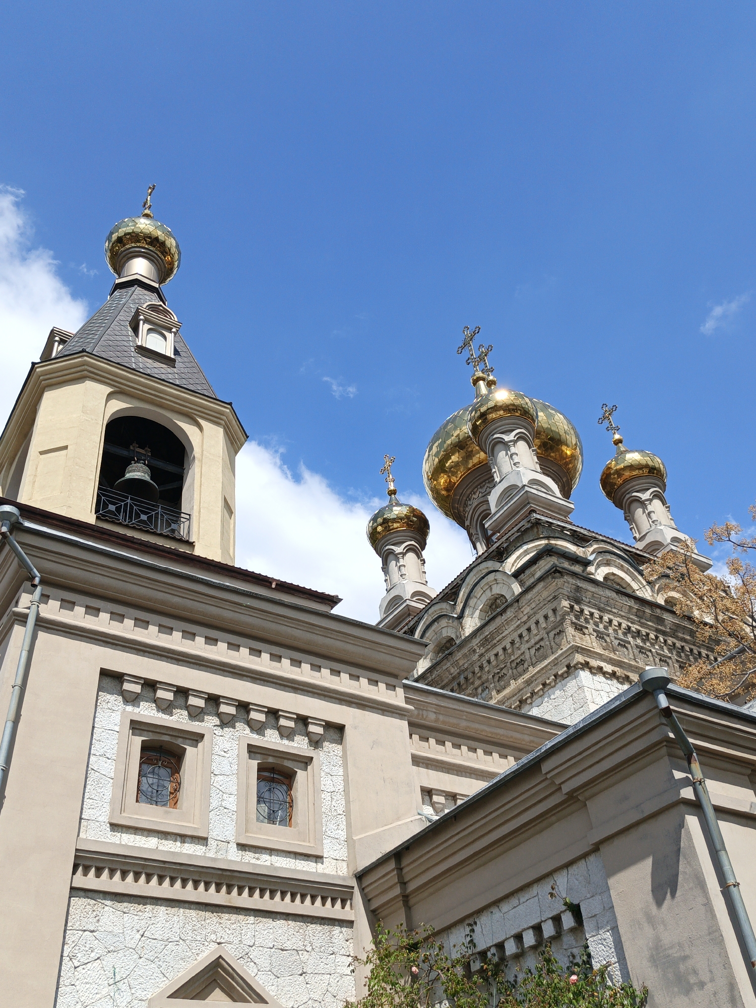 The image showcases an Eastern Orthodox church in an unidentified location. The architectural details include a bell tower with a gray roof and a golden dome, and the main structure of the church with several golden domes topped with Orthodox crosses. The walls of the building are a mix of beige plaster and white stone. The sky is blue with scattered white clouds, indicating a bright day. There are green plants visible near the base of the building.