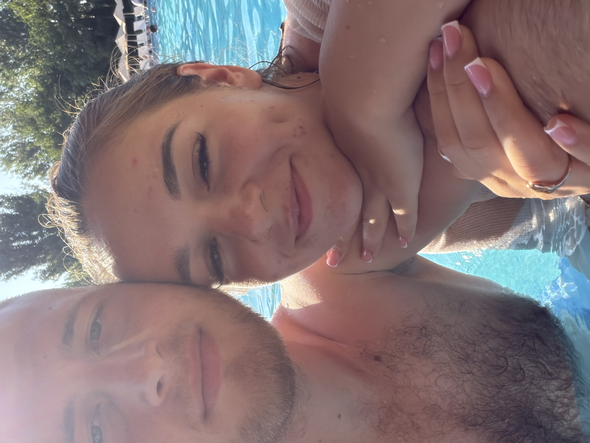 In a close-up, sun-drenched selfie taken in the swimming pool at the "Clubul de férias" resort in Sociteni, Moldova, a couple is partially submerged in the bright blue water. The man on the left, with short brown hair and stubble, faces the camera, his left shoulder and upper chest visible. To his right, a woman with her hair pulled back, appears to be resting her head on his shoulder and wrapping her arms around his chest. She is smiling, with dimples visible on her cheeks. Her hands, adorned with French manicured nails and a ring on her left hand, are intertwined with his. In the background, the shallow end of the pool is visible with lounge chairs and umbrellas, and lush green trees line the background. The sun is bright, casting a warm glow on the couple and highlighting water droplets on their skin.