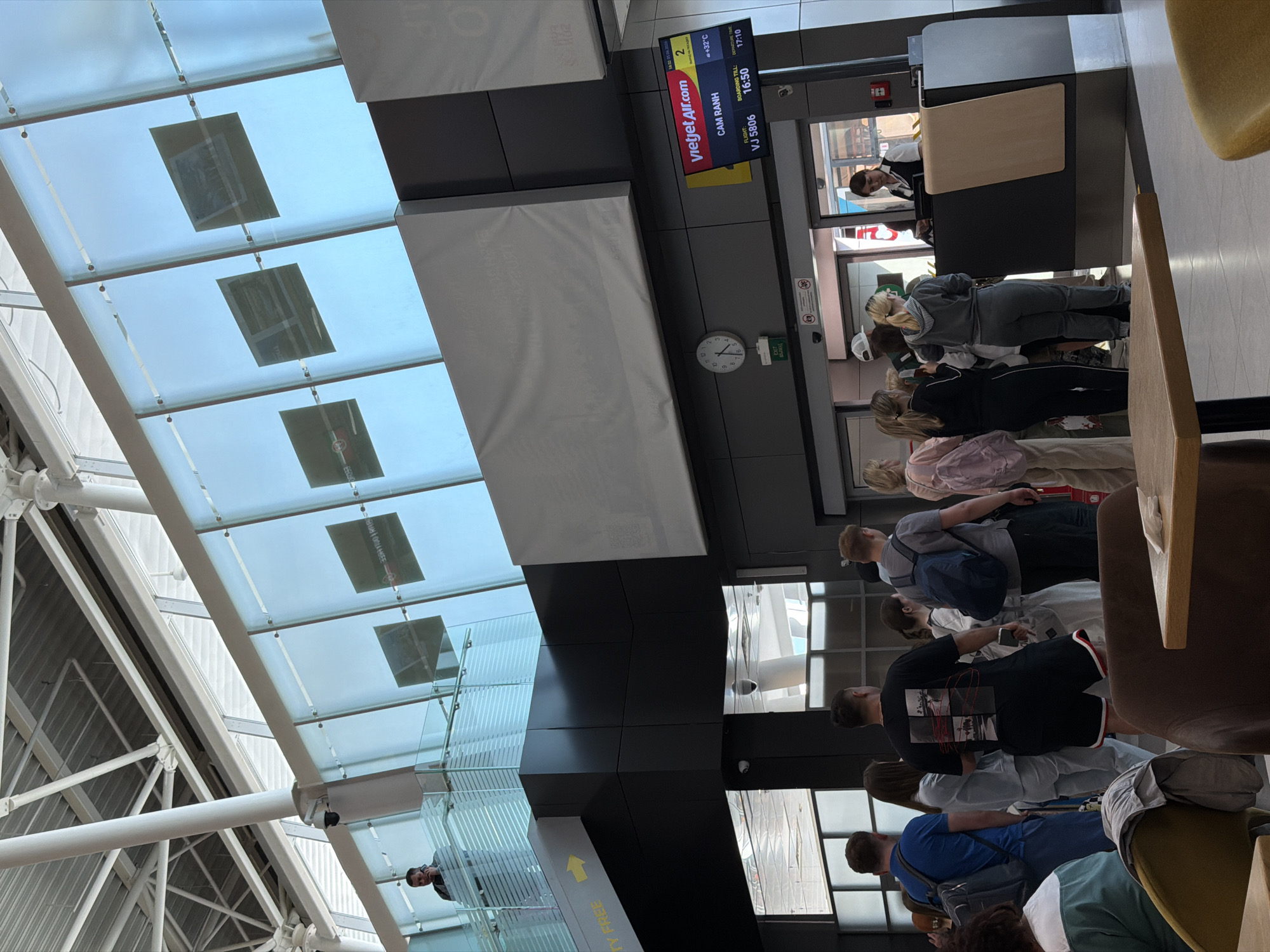At a boarding gate in an airport in Togashevo, Russia, several passengers are seen queuing, presumably awaiting their flight. The scene is captured from a slightly elevated perspective, looking down the line of people towards the boarding counter.
In the foreground, a queue of passengers stands, many with backpacks, facing a boarding counter. An airport staff member, wearing a white shirt and black waistcoat, is visible behind the counter, looking towards the queue. To the right of the counter, a flight information display screen shows details for a Vietjet Air flight. The flight, VJ 5806, is destined for Cam Ranh, Vietnam. Boarding is scheduled until 16:50, with a departure time of 17:10. The screen also indicates a temperature of +32°C. A round clock on the wall beneath the screen shows the current time to be approximately 16:25, corroborating the late afternoon setting. A green "EXIT" sign is also visible near the clock.
The airport features modern architecture with a high, slanted glass ceiling supported by white metal beams, allowing natural light into the terminal. Reflections of the internal structure and potentially external elements are visible on the glass. There are also large, white, cushion-like panels or structures suspended from the ceiling. A yellow arrow sign, partially visible, points to the right with the text "TY FREE" and "109" also indicated from OCR, suggesting proximity to a duty-free area. The atmosphere appears orderly as passengers wait.