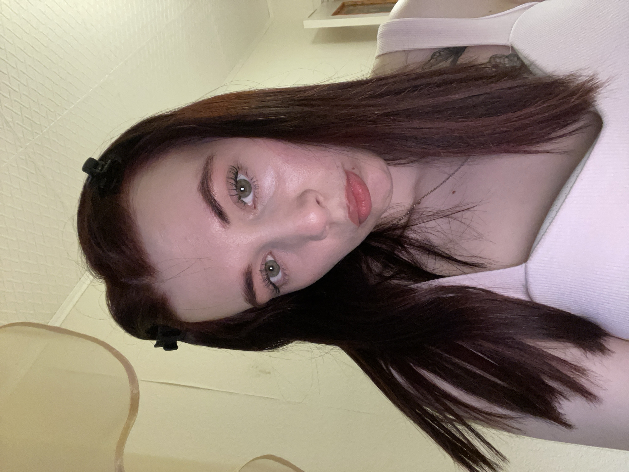 A close-up, angled portrait features a young woman with shoulder-length, dark brown hair, pulled back with black clips. She has fair skin, green eyes, and pink lips. She wears a light pink top. A partial view of a wall and a pale yellow object is visible in the background. There is no discernible indication of the location being Chaikovsky, Russia.