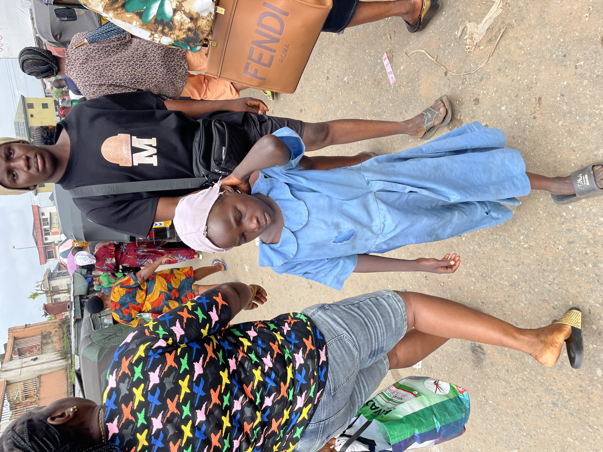 In Nkpor, Nigeria, a young girl in a light blue dress walks with her hand near her head on a gritty street. She is surrounded by people. To her left, a woman wearing a colorful, patterned top and denim shorts walks away from the camera, carrying a green shopping bag. To her right, a young man in a black t-shirt with a "M" logo and a beige beanie looks forward, with a dark crossbody bag slung over his shoulder. Behind him, a woman in a leopard-print top and orange pants walks away, and another person is visible holding a brown Fendi bag. The background features buildings, parked vehicles, and other pedestrians. The scene appears to be a busy marketplace or street.