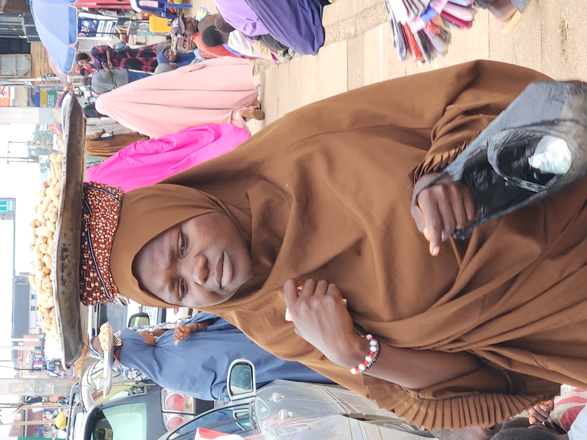 The image captures a bustling scene at a market in Jos, Nigeria. In the foreground, a woman wearing a brown hijab and a matching wrapper is the central subject. She is carrying a large tray of peanuts on her head, balanced precariously on a patterned fabric cushion. Her gaze is directed downwards and slightly to the left, giving her a contemplative or watchful expression. On her left wrist, she wears a bracelet made of red and white beads. In her right hand, she holds a black plastic bag containing some white material.
Behind her, other individuals are visible, contributing to the sense of a busy marketplace. To the left, a woman in a blue hijab is partially obscured by a silver car. Further back, several people are walking and interacting, some wearing colorful headscarves and traditional attire. Pink and pale pink fabric, possibly part of headscarves or clothing, are prominent in the middle ground, adding vibrancy to the scene. In the background, market stalls with various goods are suggested, and the overall environment appears to be outdoors, likely on a paved street or walkway. The lighting suggests it is daytime.