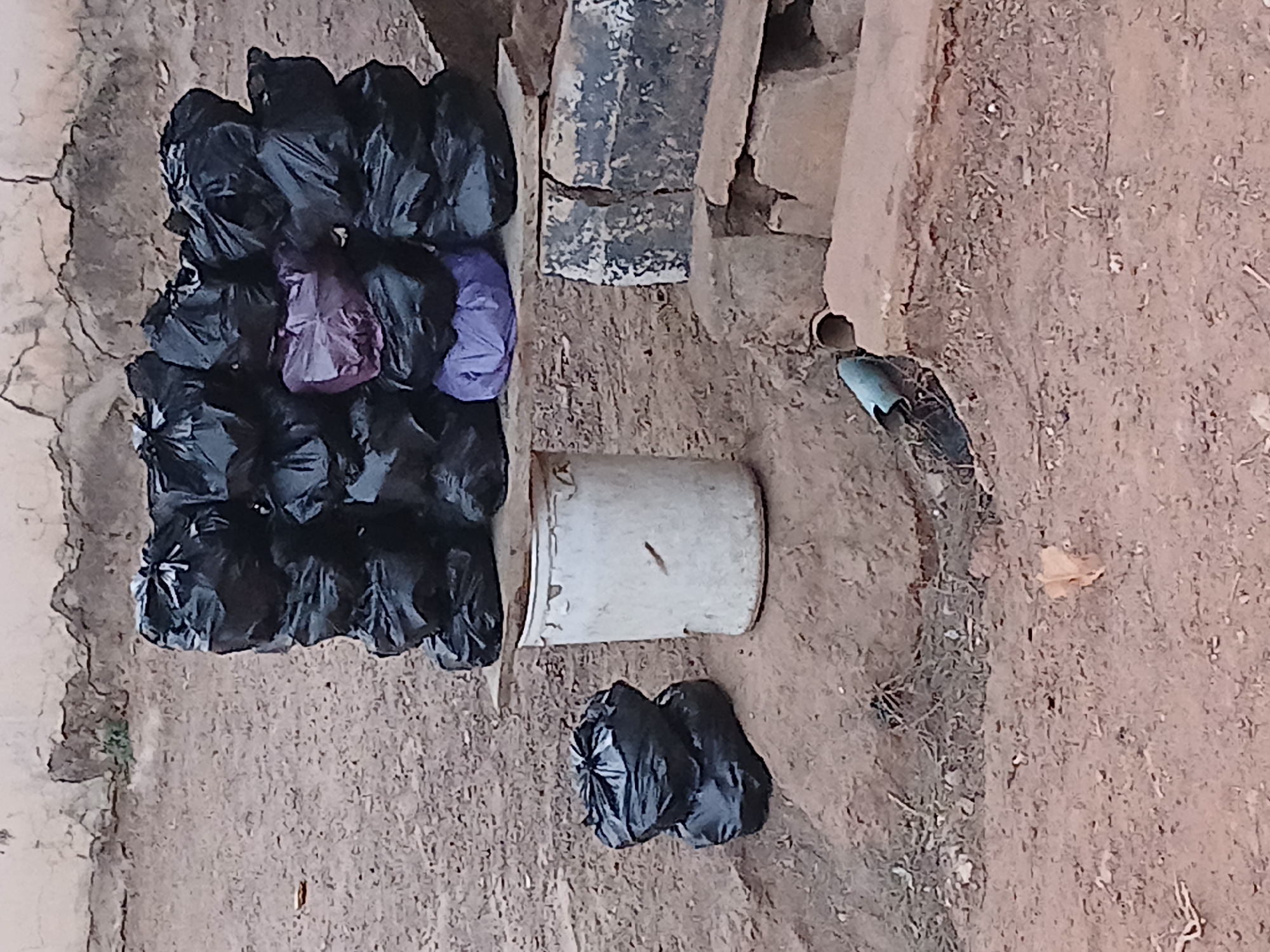 The image shows a collection of black and purple plastic bags, a white bucket, and a section of a concrete structure. The bags are grouped together in what appears to be an outdoor area in Sabon Tasha, Nigeria. The ground is earthen, and the concrete structure is broken in some places. A drainage pipe is visible near the concrete structure.