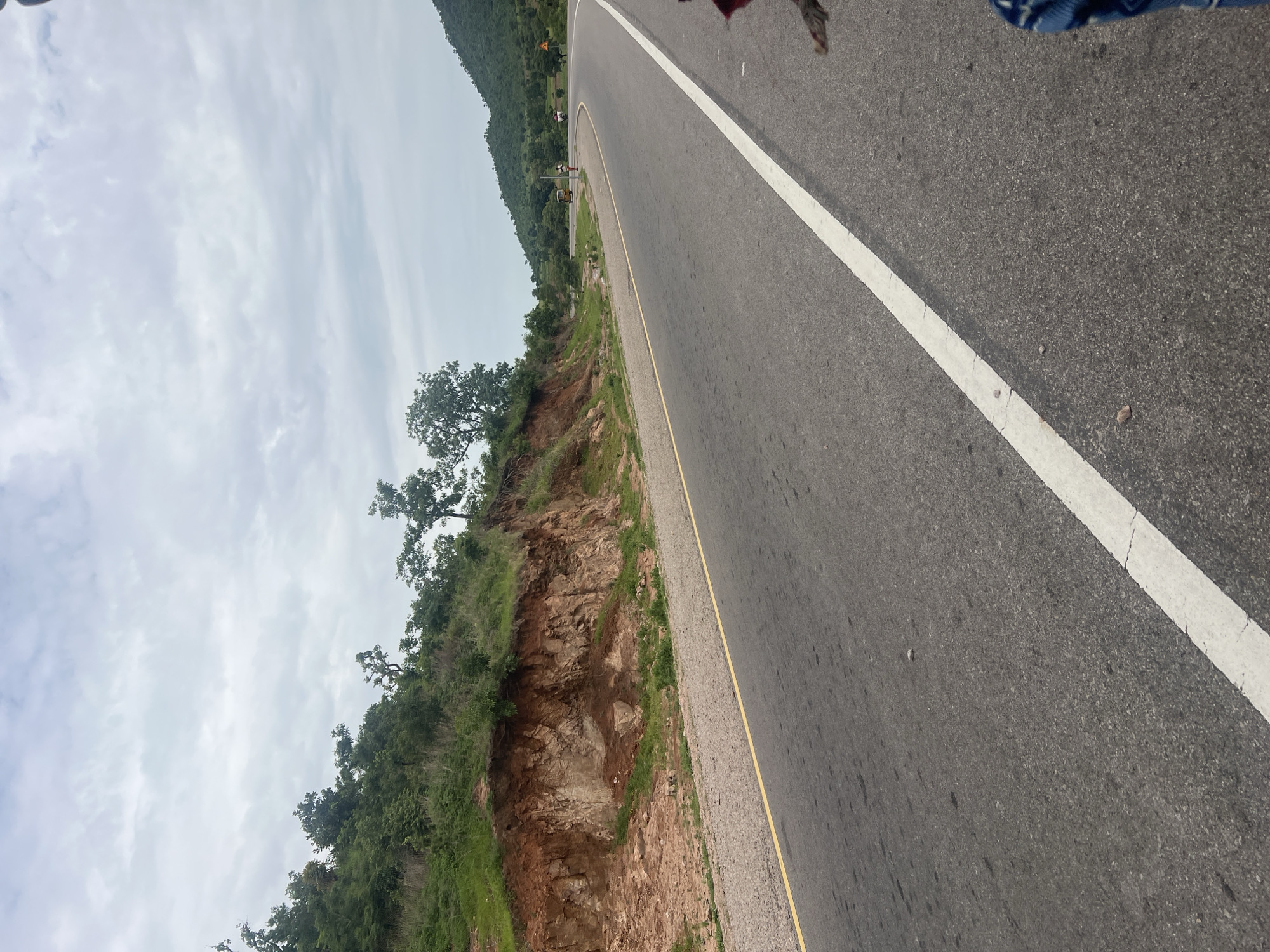 The image shows a road curving to the right, with a steep embankment on the left. The embankment is composed of reddish-brown soil and rocks, with green vegetation growing on top and along its base. The road is asphalt, with a yellow line marking the shoulder on the left and a white line on the right lane. In the distance, the road passes by rolling green hills, some of which are covered in trees. There are a few small buildings visible in the distance, as well as some signs of human activity, including a yellow vehicle and two people on the side of the road. The sky is overcast with a gray, cloudy sky. The scene depicts a rural or semi-rural road in Minda, Nigeria.