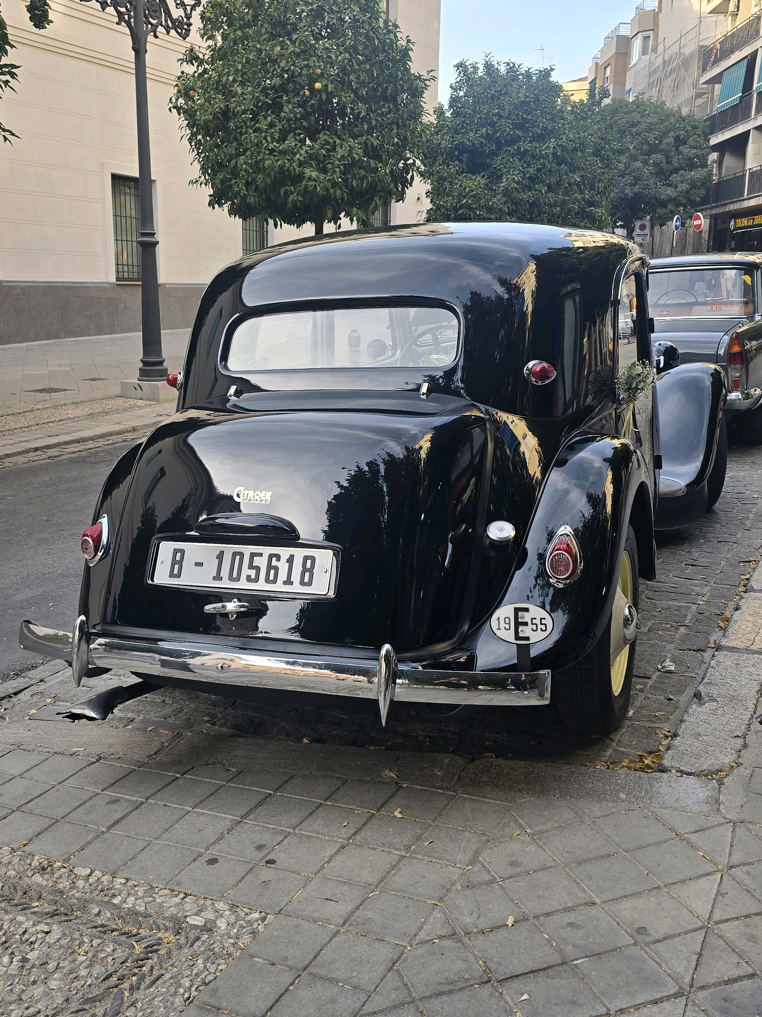 The image captures the rear of a vintage black Citroën car parked on a cobblestone street in Granada, Spain. The car has a chrome bumper, red taillights, and a license plate that reads "B-105618". A circular sticker with "1955" is also visible on the rear fender. The street is lined with trees and buildings, suggesting an urban setting. The car appears to be part of a procession or event, as indicated by a small bouquet of flowers on the passenger side rear fender. The overall lighting suggests it is daytime.