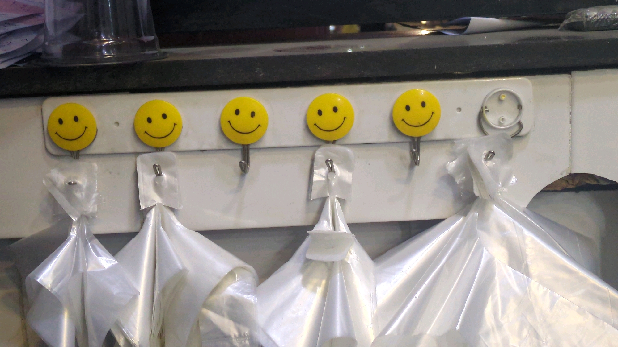 The image shows five yellow smiley face hooks mounted on a white wall. Each hook has a small, bent metal piece extending downwards. Four of the hooks are holding white plastic bags, which are bunched up at the top. The fifth hook, which is empty, is located between the second and third smiley face from the left, and it has a small, empty ring attached to it. The smiley faces are round with black eyes and mouths. The background wall is white and slightly textured. Above the hooks, a dark countertop or shelf is visible, with some blurry items on it. The lighting appears to be overhead, casting shadows on the bags. No people are visible in the image. The scene is indoors. No specific location in Lahore, Pakistan can be determined from the image.