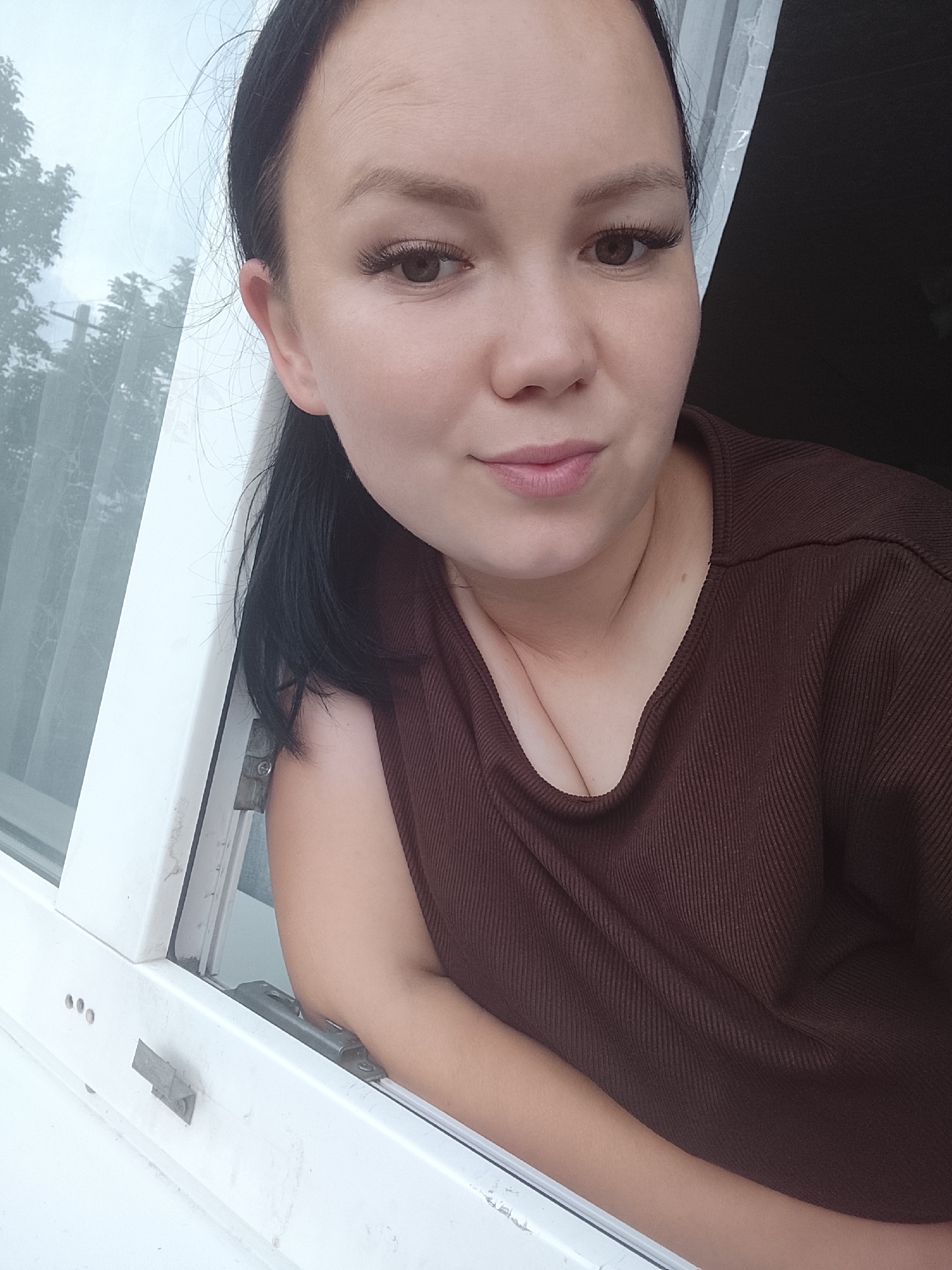 A woman with dark hair and false eyelashes is leaning out of a white window frame, looking at the camera. She is wearing a brown, ribbed, V-neck top. Her right arm is resting on the windowsill. Outside the window, trees are visible through a translucent curtain or blind. The background on the right is dark. The scene appears to be in Kuyeda, Russia, based on contextual information not present in the image itself.