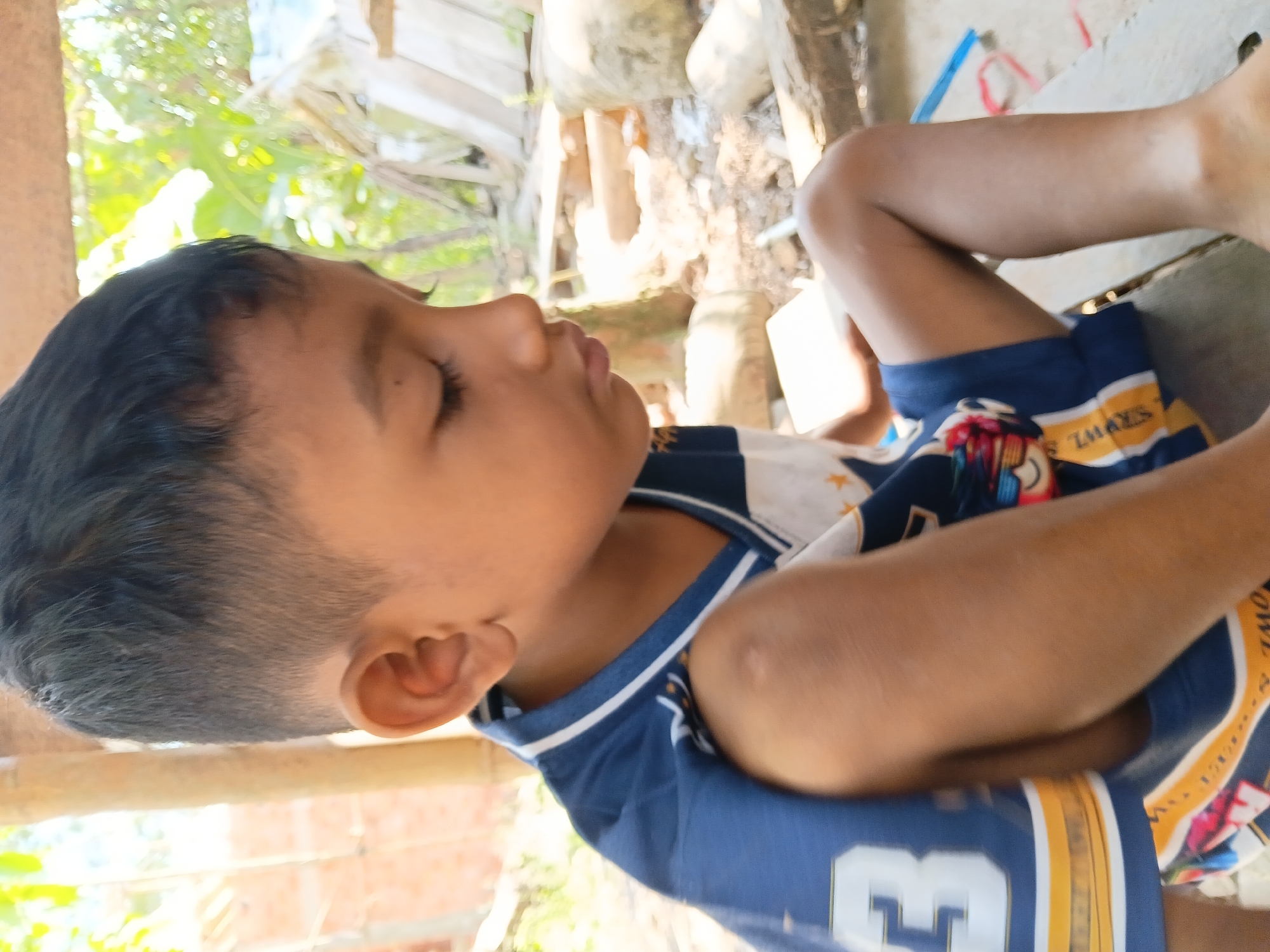 A young boy, with dark hair and brown skin, is shown in a medium close-up shot, looking downwards and to his right. He is wearing a blue jersey with white and yellow stripes, and a number "3" is visible on the left side of his chest. The boy is seated on a wooden surface, with his legs crossed. His right leg is extended, and his bare foot is visible. The background is blurred, but appears to be an outdoor environment, possibly a rural area in Margo Rukun, Indonesia, with some foliage, wooden structures, and rocks. The lighting suggests it is daytime, with sunlight filtering through.