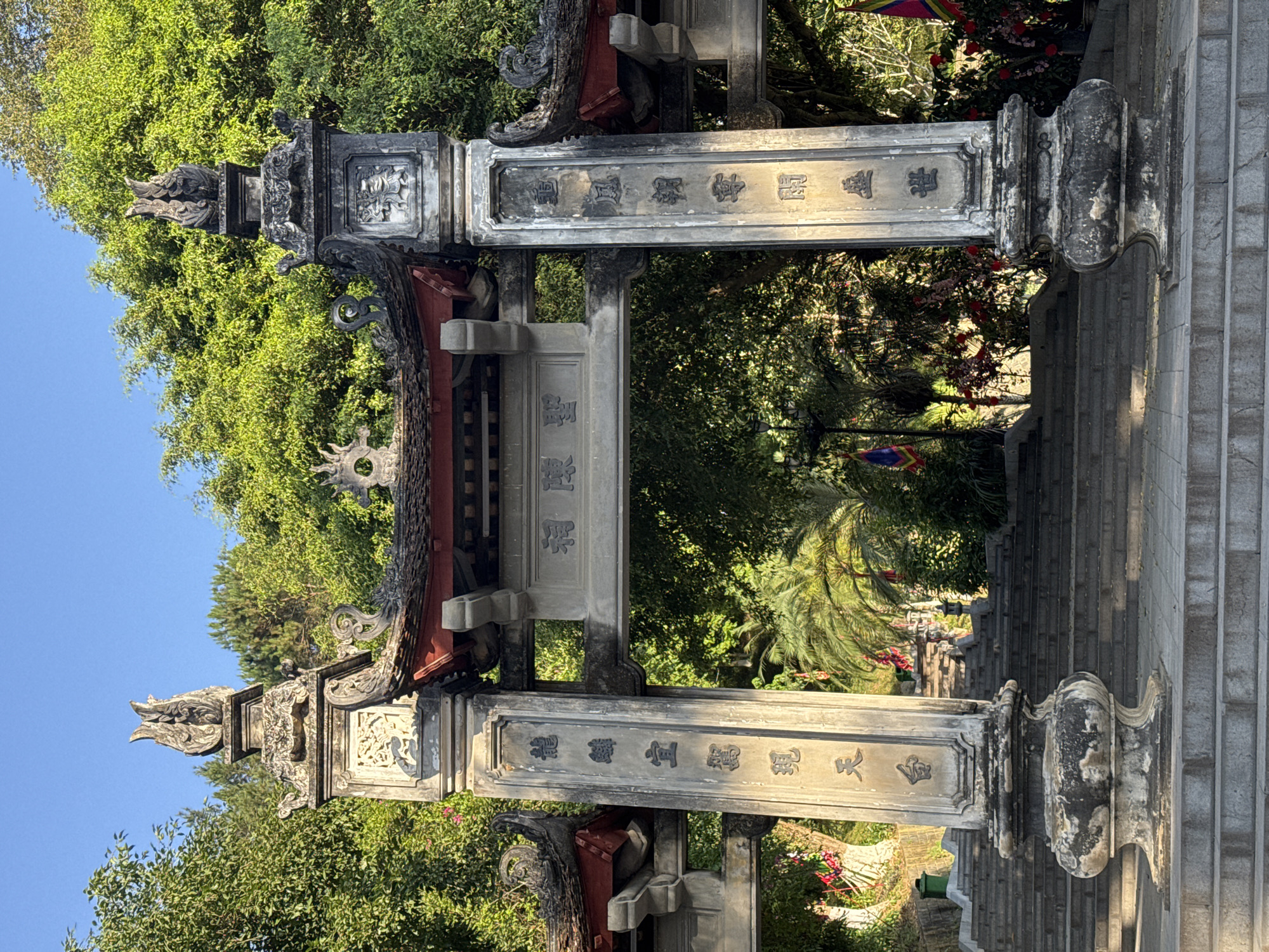 The image captures an ornate stone gate structure, likely an entrance to a temple or historical site, located in Lao Cai Ward, Vietnam. The gate is constructed of gray stone, adorned with traditional Vietnamese architectural features such as intricate carvings, curved roof elements, and dragon-like figures atop the pillars. Inscriptions in Vietnamese characters are visible on the stone surfaces of the gate. The gate opens to a pathway or staircase leading upwards, surrounded by lush green trees and foliage. The sky above is blue, suggesting a clear day. The overall scene conveys a sense of cultural heritage and serene natural surroundings.