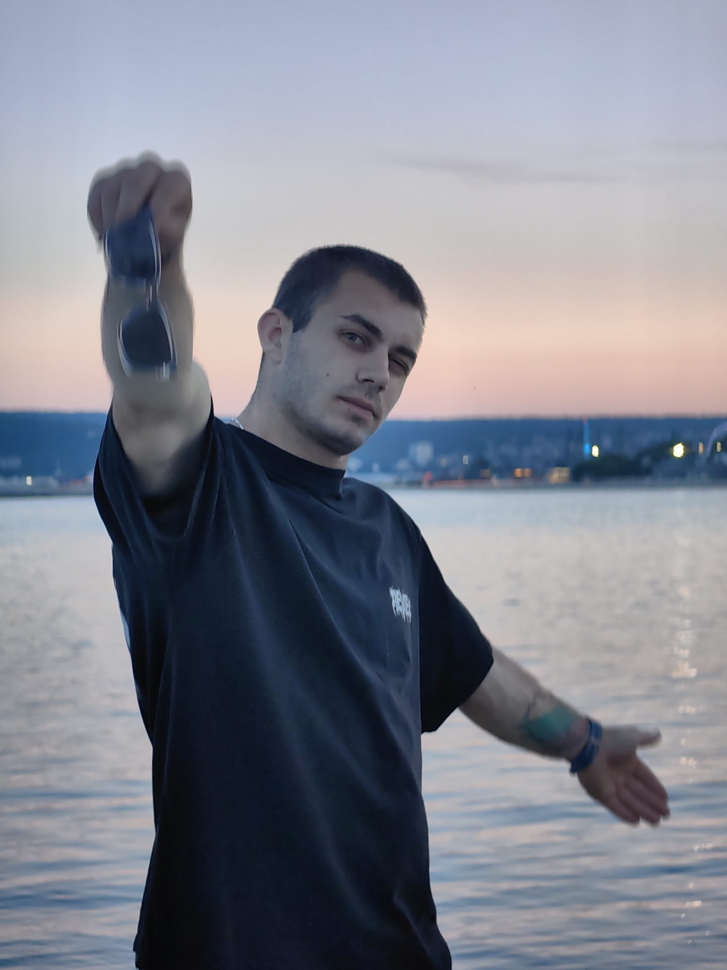 A young man stands on the edge of a body of water during twilight. He is wearing a black t-shirt and holding a pair of sunglasses in his right hand, extended towards the camera. His left arm is outstretched, palm facing forward. The background features a city skyline with lights reflecting on the water. The sky is a gradient of blue and pink. The man has short dark hair and appears to be looking at the camera with a serious expression.
