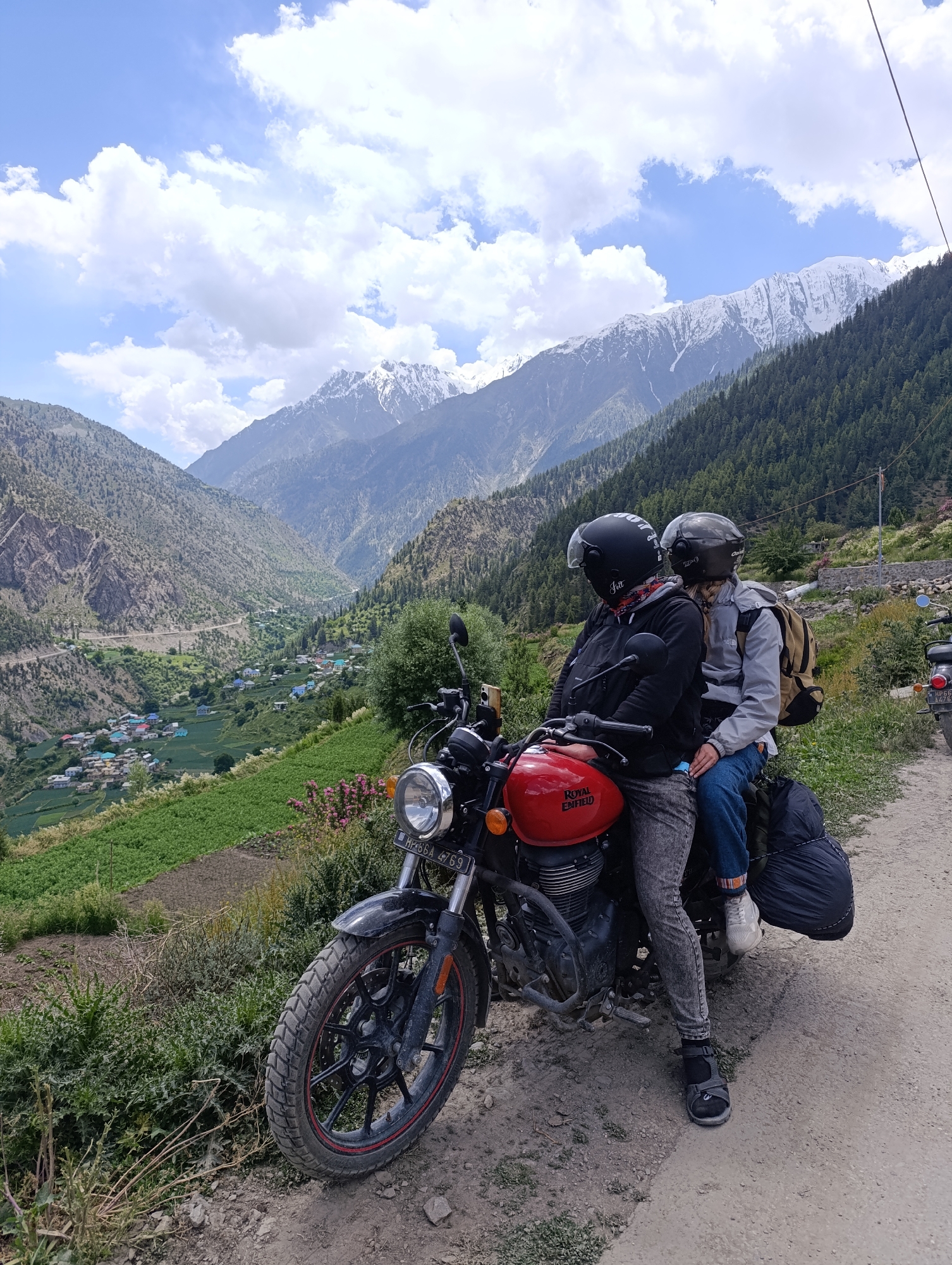 Two individuals wearing helmets are seated on a red Royal Enfield motorcycle. The person in the front is wearing a black jacket, grey patterned pants, and a black helmet with a red bandana visible underneath. They are holding the handlebars. The person in the back is wearing a grey jacket, blue jeans, and a silver helmet. They have a beige backpack on their back and their arm is around the person in front. Luggage is secured to the rear of the motorcycle.
The motorcycle is parked on a dirt road overlooking a valley. In the valley, there is a village with colorful houses nestled amongst green fields. A winding road is visible leading into the valley. The background features steep, tree-covered mountains, with snow-capped peaks in the distance. The sky is blue with scattered white clouds. The scene is set in the mountainous region of Kishorl, India.