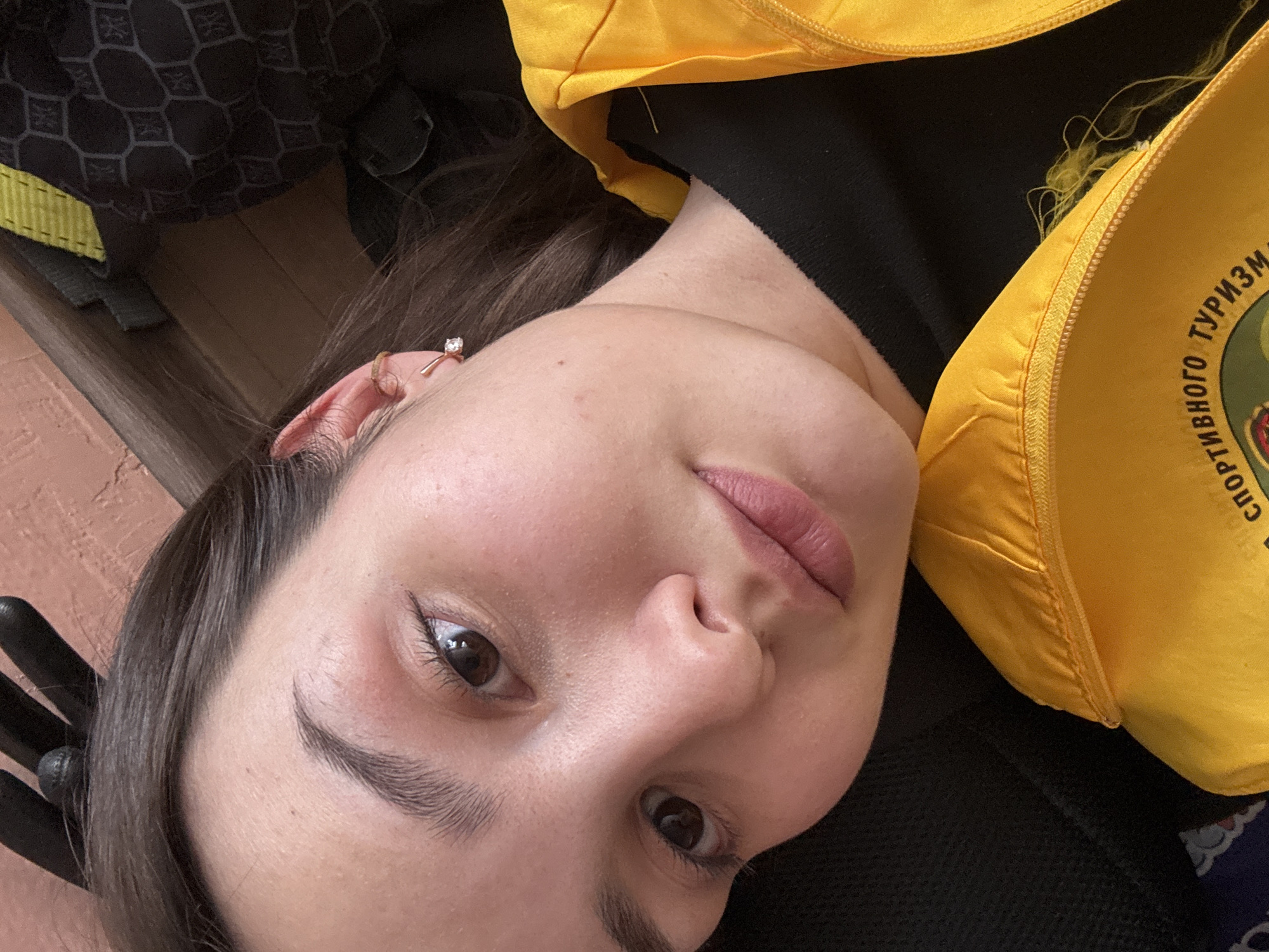 A close-up, angled portrait depicts a female subject, encompassing her face, neck, and upper chest, in an indoor setting. The subject has dark brown hair, dark brown eyes, and fair skin. Her right ear is visible, adorned with a small hoop earring near the helix and a single-stone stud earring on the lobe. Her lips are closed, exhibiting a pink hue. She wears a black garment beneath a bright yellow zippered outer garment, visible in the lower right quadrant. The yellow garment features partially visible embroidered Cyrillic text, reading "СПОРТИВНОГО ТУРИЗМ" (SPORT TOURISM). Loose yellow threads are apparent near the zipper of the yellow garment. The background consists of an out-of-focus reddish-brown textured surface on the upper left and a dark object with a geometric pattern and a yellow strap on the upper right, consistent with a backpack or similar item. The subject maintains direct eye contact with the camera. The image itself contains no discernible features to confirm the specific location of Tayshet, Russia.