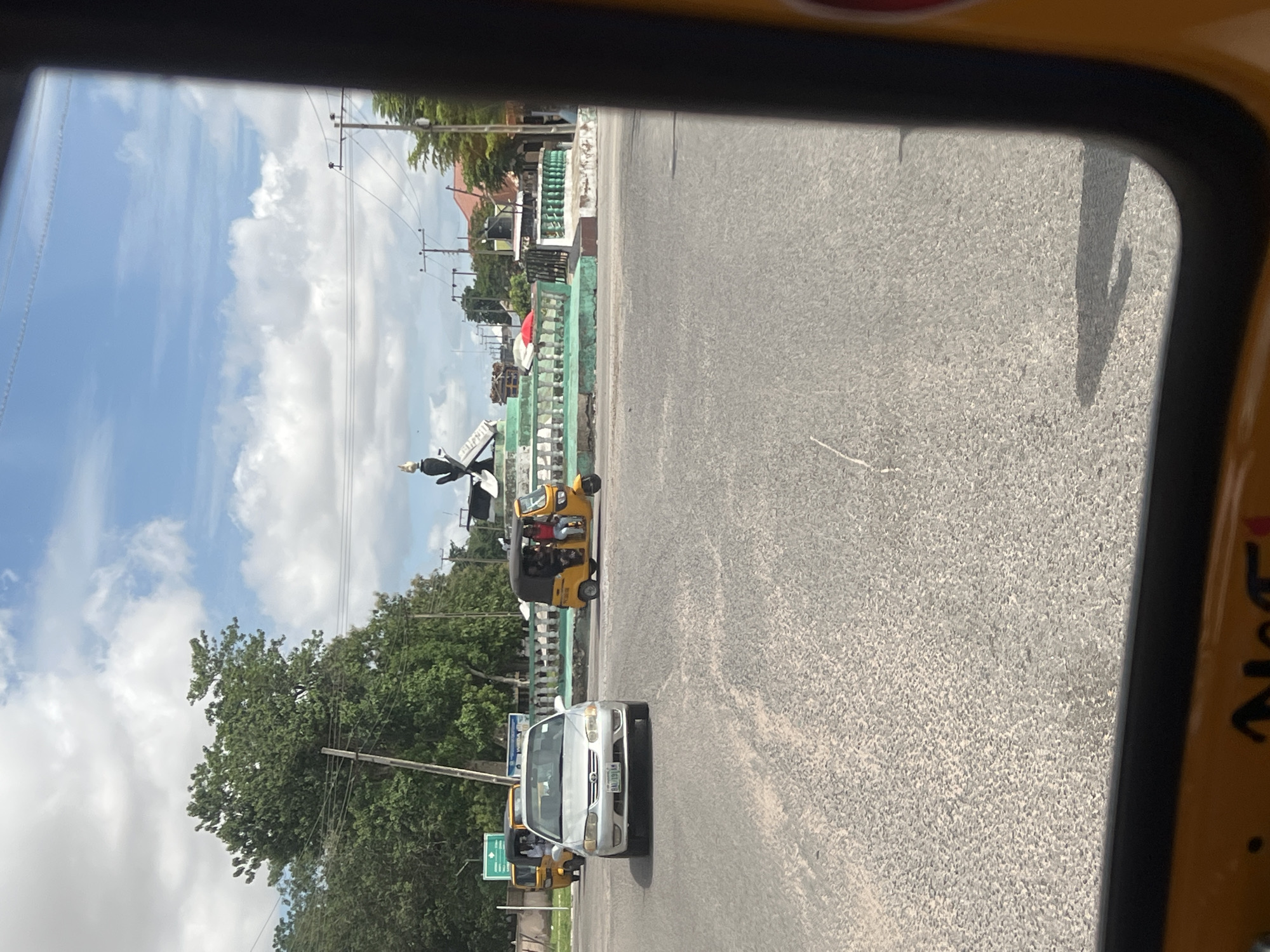 This image captures a street scene at a roundabout in Jalingo, Taraba State, Nigeria, specifically identifiable as a significant intersection, possibly the Taraba State Welcome Gate or a similar prominent roundabout.
In the center of the roundabout stands a monument featuring a dark, stylized sculpture, resembling an eagle or a similar large bird, perched atop a pedestal with an open book motif. The monument's base is adorned with green and white balustrades, indicating some wear and local architectural style. Behind the monument, a red umbrella is visible, along with distant buildings, one having a reddish roof.
A silver sedan, bearing the Nigerian license plate "BAL191AW", is positioned in the foreground, facing the camera. To its left, partially obscured, is a yellow tricycle (keke Napep). Another yellow tricycle, with passengers visible inside (one in a red top), is positioned to the right of the silver car, closer to the roundabout. A third yellow tricycle is visible further in the background to the left.
The surrounding environment features abundant green trees, several utility poles with numerous power lines crisscrossing the sky, and a partly cloudy blue sky. The road is asphalt, appearing dry, with visible shadows indicating daylight conditions. The photograph is taken from inside a vehicle, likely another yellow tricycle, whose yellow frame and partial lettering "N" are visible in the bottom left corner, and its shadow is cast on the road in the bottom right.