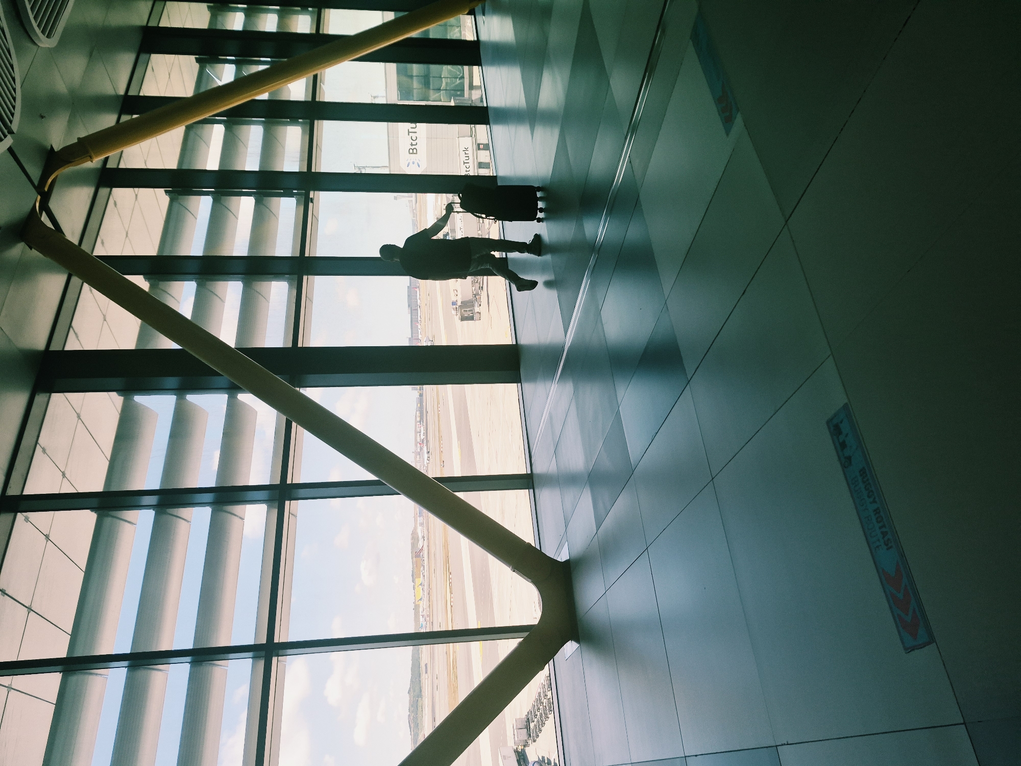 In the city of Arnavutköy, Turkey, a solitary figure walks through a modern airport terminal, their silhouette cast against a large window overlooking an airfield. The individual, clad in a dark t-shirt and jeans, is pulling a dark rolling suitcase. The terminal's architecture is characterized by large, expansive windows with horizontal louvers that filter the daylight, and prominent yellow structural beams that support the ceiling. The floor is tiled in a grid pattern, with subtle directional markers visible. Outside, an airport tarmac is visible, with several aircraft and airport support vehicles. The scene suggests a moment of travel, with the person likely heading to a departure gate or arriving from a flight.