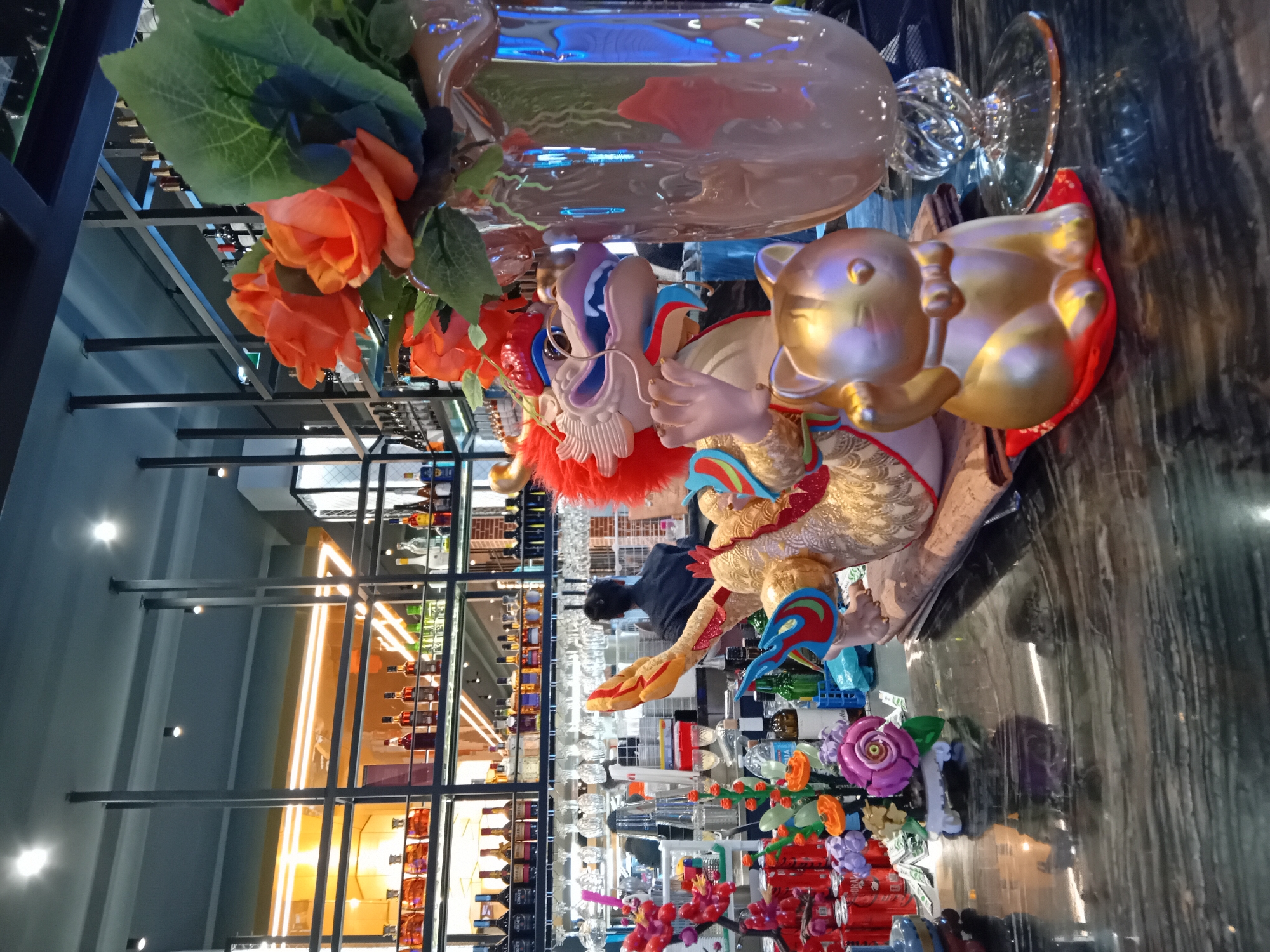 The image displays an indoor scene, likely a bar, lounge, or liquor store, viewed from a slightly elevated perspective. In the immediate foreground, on a glossy dark counter that reflects the artificial lighting, is a vibrant and ornate decorative statue. This statue features a colorful, stylized head resembling a mythical creature or a lion dance mask, with a prominent red fluffy mane, a blue and white face, and a golden, intricately textured body adorned with red and blue accents. It holds a smaller, golden, pig-like figurine. To the statue's left, a large, clear glass vase contains artificial orange roses and green leaves. Another golden figurine, possibly a Maneki-neko or lucky cat, rests on a red cushion beside the main statue.
The background is dominated by extensive shelving units constructed from dark metal, showcasing numerous bottles of what appears to be alcohol. Various clear drinking glasses, including stemware, are neatly arranged on these shelves and counter surfaces. A single person, seen from behind and wearing a dark shirt, is visible in the mid-ground, appearing to be working or attending to the bar area.
Further details in the background include more colorful artificial flowers, such as purple and orange roses, red berries, and stacks of red beverage cans. The lighting is artificial, with warm yellow light strips illuminating the shelves and cool white spotlights visible overhead. Reflections from these lights are prominent on the glossy surfaces and glassware. No readable text is visible on any of the bottles or signs. The scene conveys a modern and well-stocked commercial establishment, possibly with an Asian-inspired decorative theme.