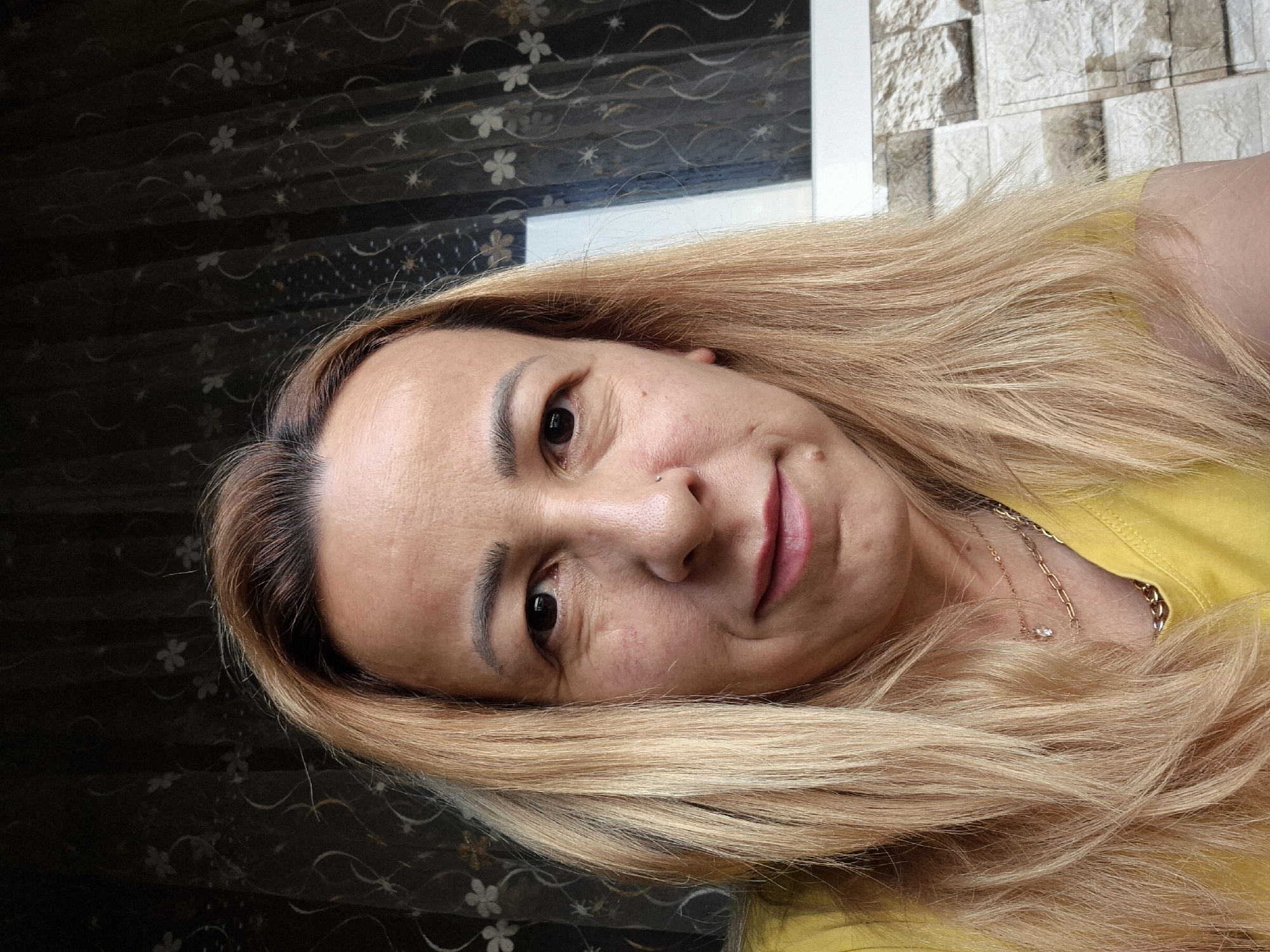 A close-up selfie shows a person lying down, with their head tilted to the left. The person has light blonde hair, which is styled straight and falls around their face. They are wearing a yellow top and a delicate gold necklace with a small pendant. The background is a blend of a dark, patterned curtain with floral designs and a textured wall that appears to be made of light-colored stone or brick. The lighting suggests it is daytime, possibly indoors given the curtains and wall textures. The person's expression is neutral, with a slight smile. There are no visible text elements in the image.