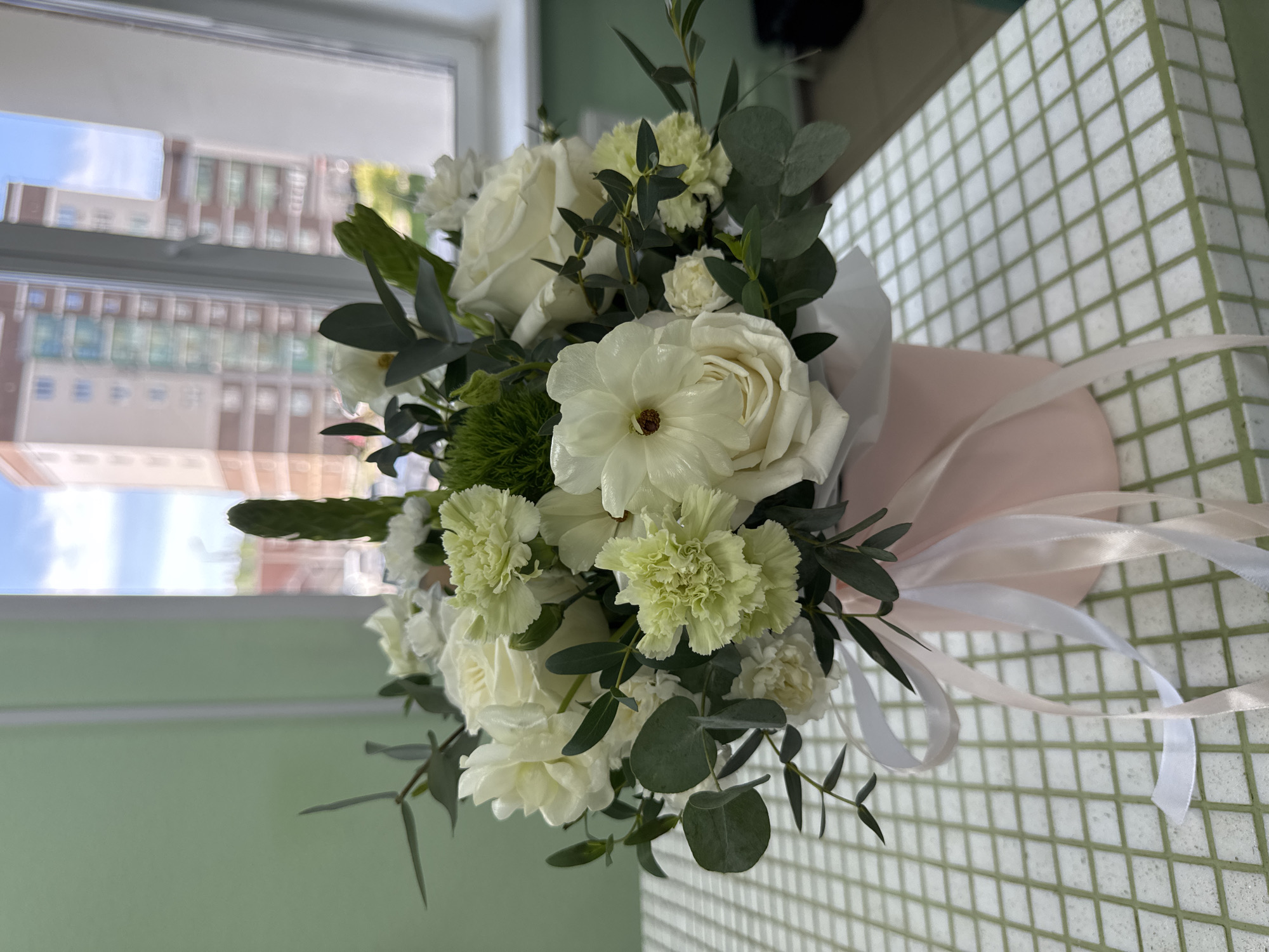 The image shows a bouquet of white roses and other white and light green flowers, arranged with eucalyptus leaves, in a pale pink wrapping. The bouquet is tied with white ribbons and is placed on a surface with a green and white checkered pattern. In the background, a window reveals a view of buildings, suggesting an urban setting. The location, Izhevsk, Russia, is indicated as context. There are no people visible in the image.