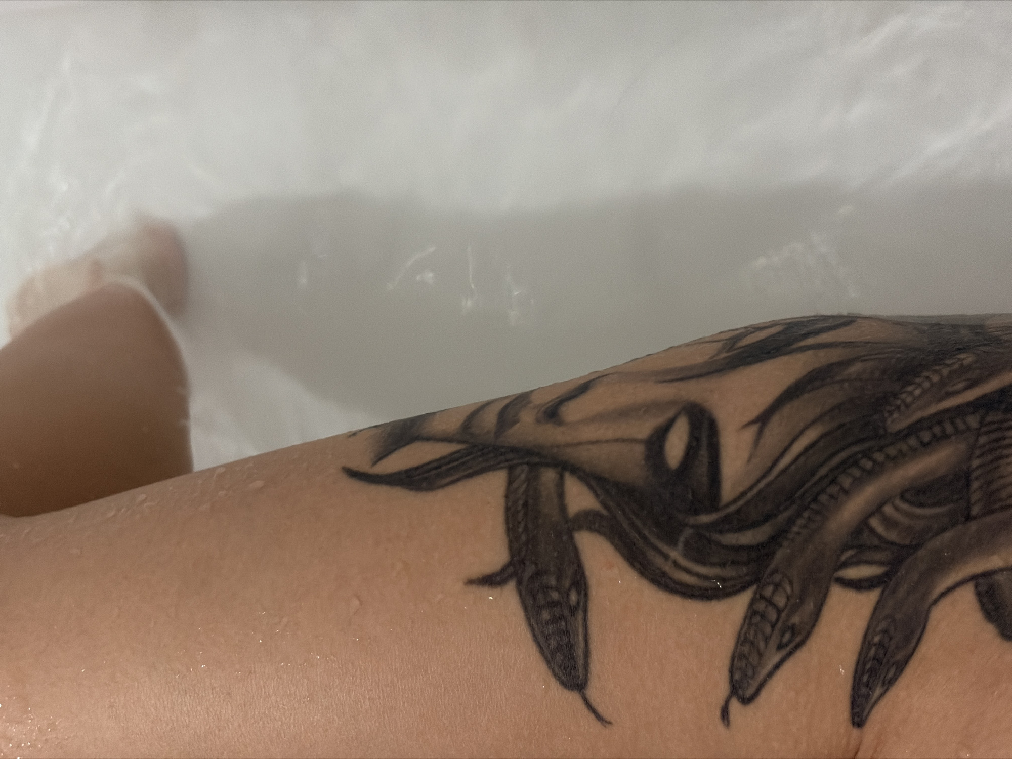 The image displays a close-up view of a person's arm and partially submerged leg, taken from a downward perspective, presumably while bathing in a bathtub in Saint Petersburg, Russia. The main subject is the person's right arm, prominently featuring a large, detailed black and grey tattoo on the upper arm or shoulder area. This tattoo depicts multiple snake heads, complete with scales and slender, forked tongues, suggesting a mythological theme, possibly Medusa.
Water droplets are visible on the skin and across the surface of the tattoo, indicating the arm is wet. A blurred section of a leg or foot is discernible in the upper left of the frame, partially submerged in the opaque, whitish bathwater, which shows some ripples or foam. The scene takes place indoors, likely in a bathroom, with soft, ambient lighting. The activity depicted is bathing or soaking in the tub. There is no visible text in the image.