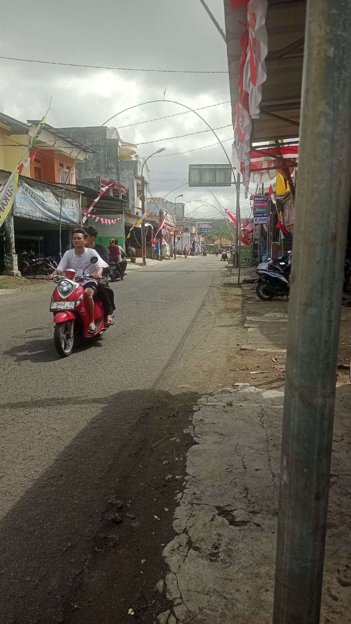 In the city of Cikalong, Indonesia, a red scooter with two male occupants, one driving and the other as a passenger, travels along a paved road. The driver, wearing a white t-shirt and dark shorts, is positioned in the front, while the passenger, in dark clothing, sits behind him. The scooter has a license plate with the characters "Z 5442 RL". The road is bordered on the right by a cracked concrete sidewalk, a metal pole, and several shops. Red and white flags, likely national flags, are displayed along the street, suggesting a celebratory event or holiday. The buildings appear to be shops with awnings and some wear nationalistic banners. The sky is overcast, with dramatic cloud formations. The general atmosphere suggests a community setting, possibly a market or a street scene in a small Indonesian town.