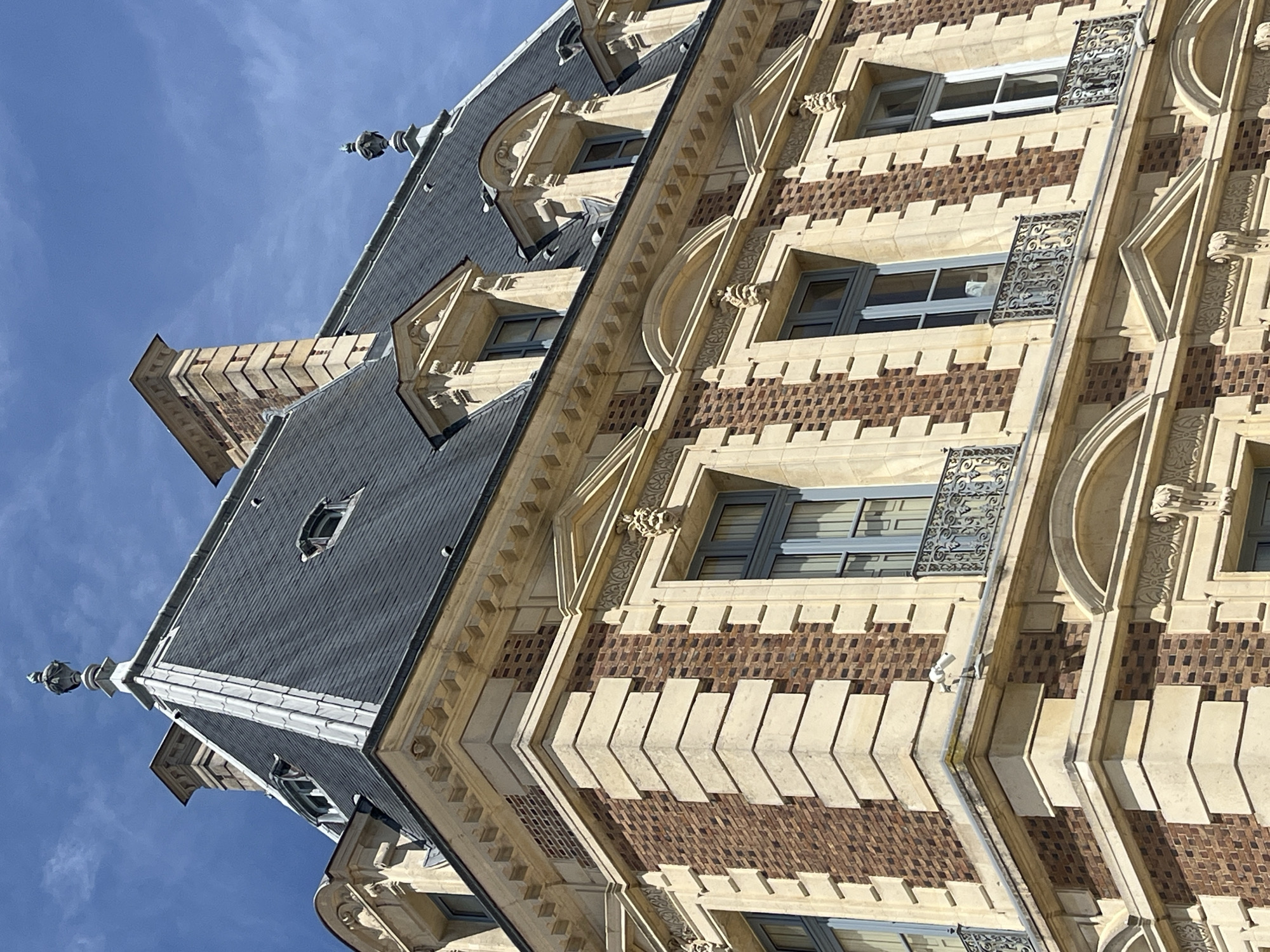 The image shows a close-up, exterior, eye-level view of a building facade in the city of Sceaux, France. The building has a mansard roof made of dark grey shingles, with several dormer windows. The facade is constructed of red brick with stone accents and decorative elements. Several windows with grey frames and decorative iron balconies are visible. The sky above is bright blue with a few wispy clouds. A security camera is mounted on a ledge near the corner of the building.