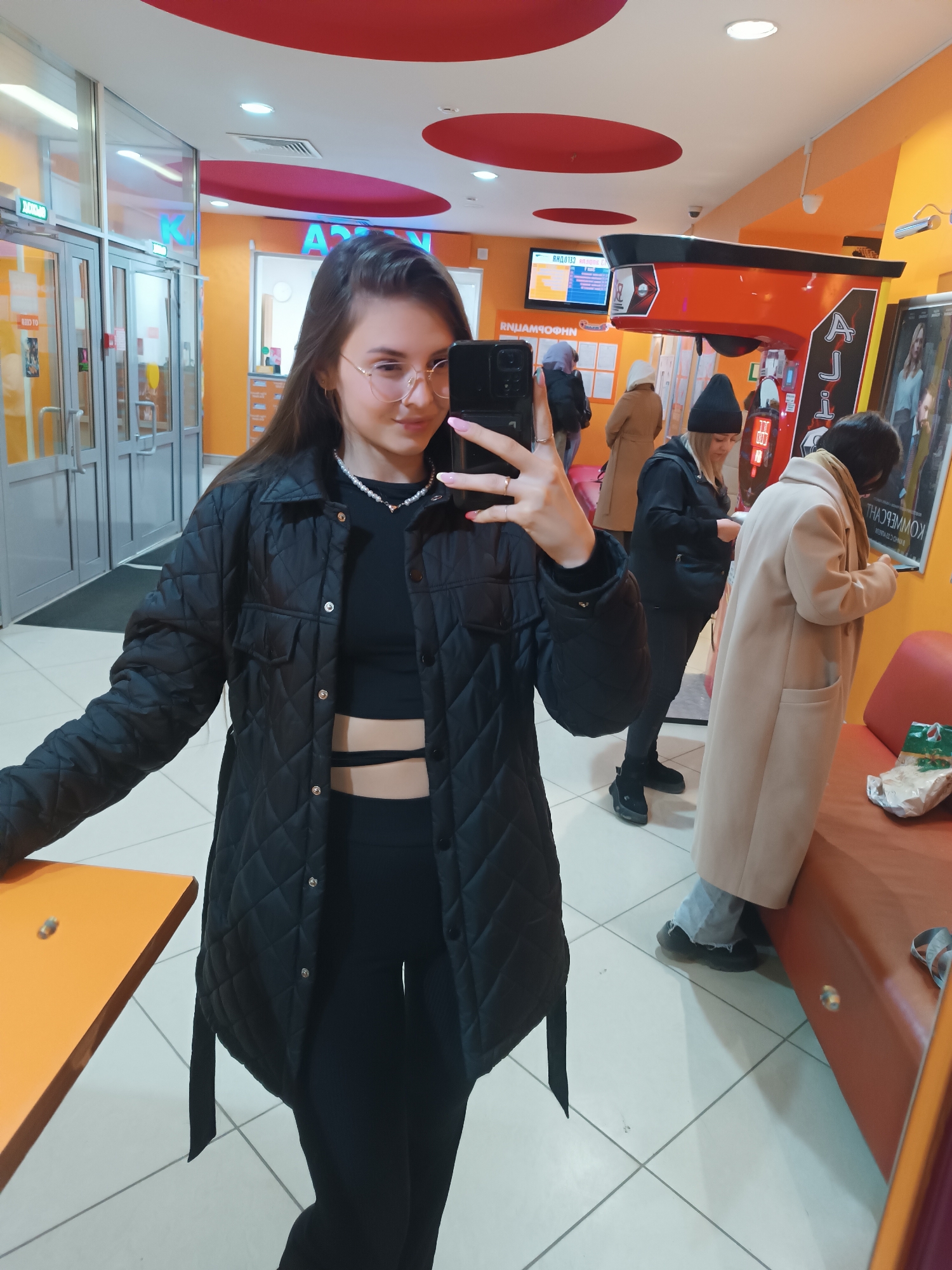 A young woman takes a selfie in a public space, likely a mall or shopping center. She is wearing a black quilted jacket, a black crop top with cutouts, and black leggings. She also wears glasses and a pearl necklace. She holds a smartphone in her right hand to take the picture.
In the background, there are several other people. To her right are two individuals; one is wearing a tan coat and looking at their phone. Further to the left is another person in a black beanie and jacket.
The environment includes commercial displays and seating areas. Orange and red circular accents are seen on the ceiling. The floor is tiled. A set of glass doors is visible in the distance. There's a possible game machine visible and an orange table next to the subject. The location is Unknown, Unknown.
