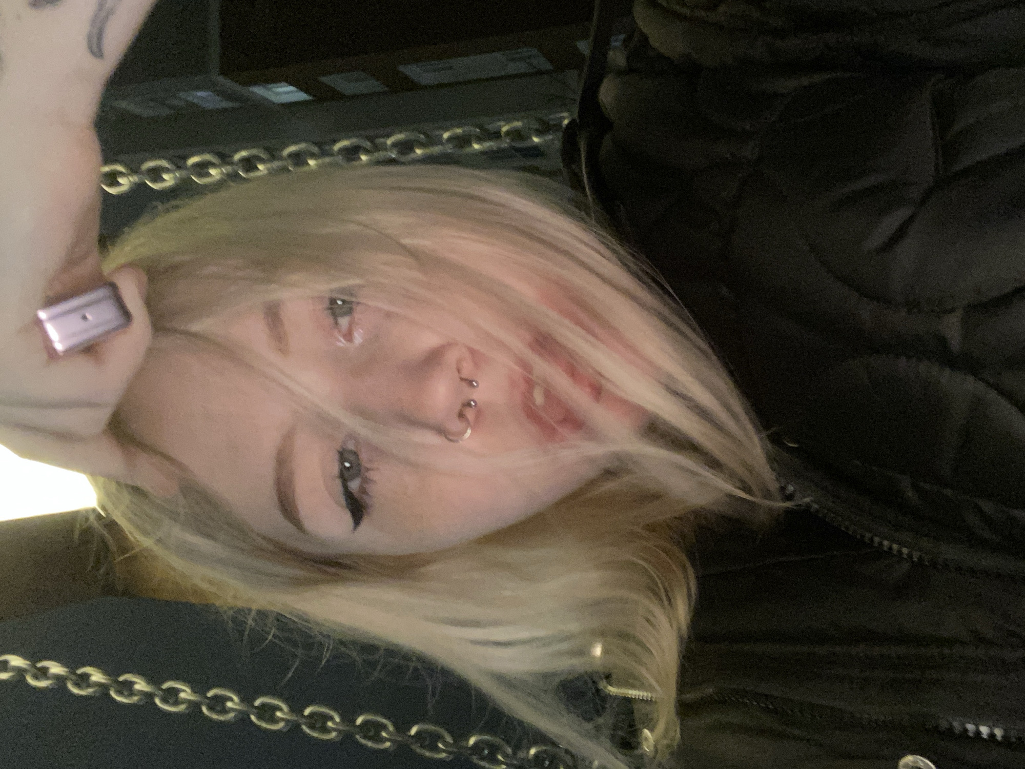 A close-up, slightly distorted selfie of a fair-skinned person with dyed blonde hair. The person has visible piercings on their nose and dark eyeliner. They are wearing a dark, possibly quilted, jacket with a visible zipper. A silver chain is visible along the lower edge of the frame. The person is holding a rectangular, possibly metallic, object in their left hand. The background is indistinct and dark. No information indicates location in Pervomaysky, Russia.