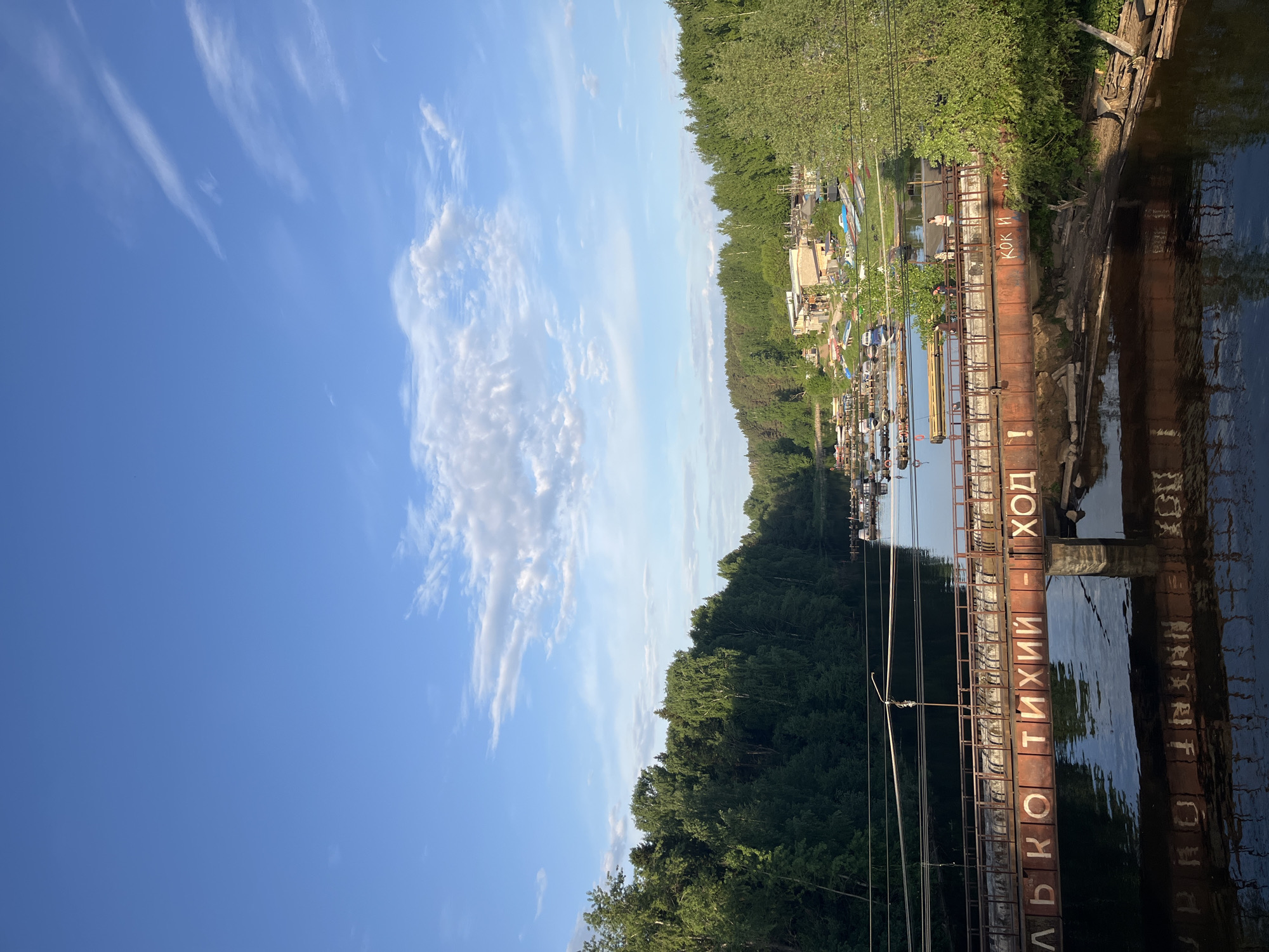 The image shows a view of a river or canal in Chaikovsky, Russia. The scene is framed by a metal structure with Cyrillic text on the right, and trees lining the riverbank on the left. The sky is blue with scattered white clouds. In the background, there are buildings and boats along the waterway. The metal structure with Cyrillic text is reflected in the water.