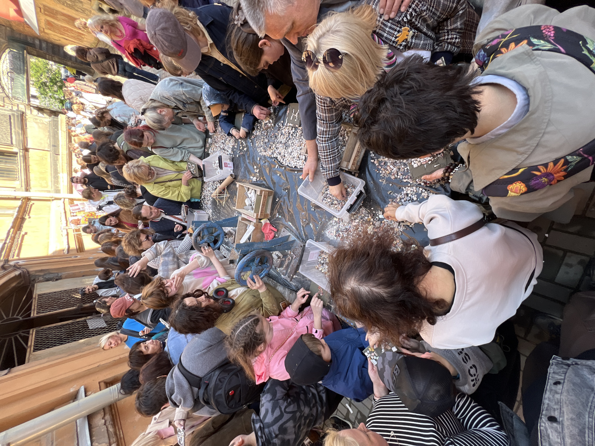 A densely packed crowd of people are participating in an activity centered around a black surface covered in white and gray mosaic pieces. They are picking up and inspecting these small fragments. Some individuals hold clear plastic containers filled with mosaics. Various wooden boxes and other objects, including at least two blue valve wheels, are scattered across the surface. The environment appears to be an outdoor alley or narrow street in Saint Petersburg, Russia, based on the architectural style. The angle of the image suggests it was taken from an elevated position, looking down on the scene.