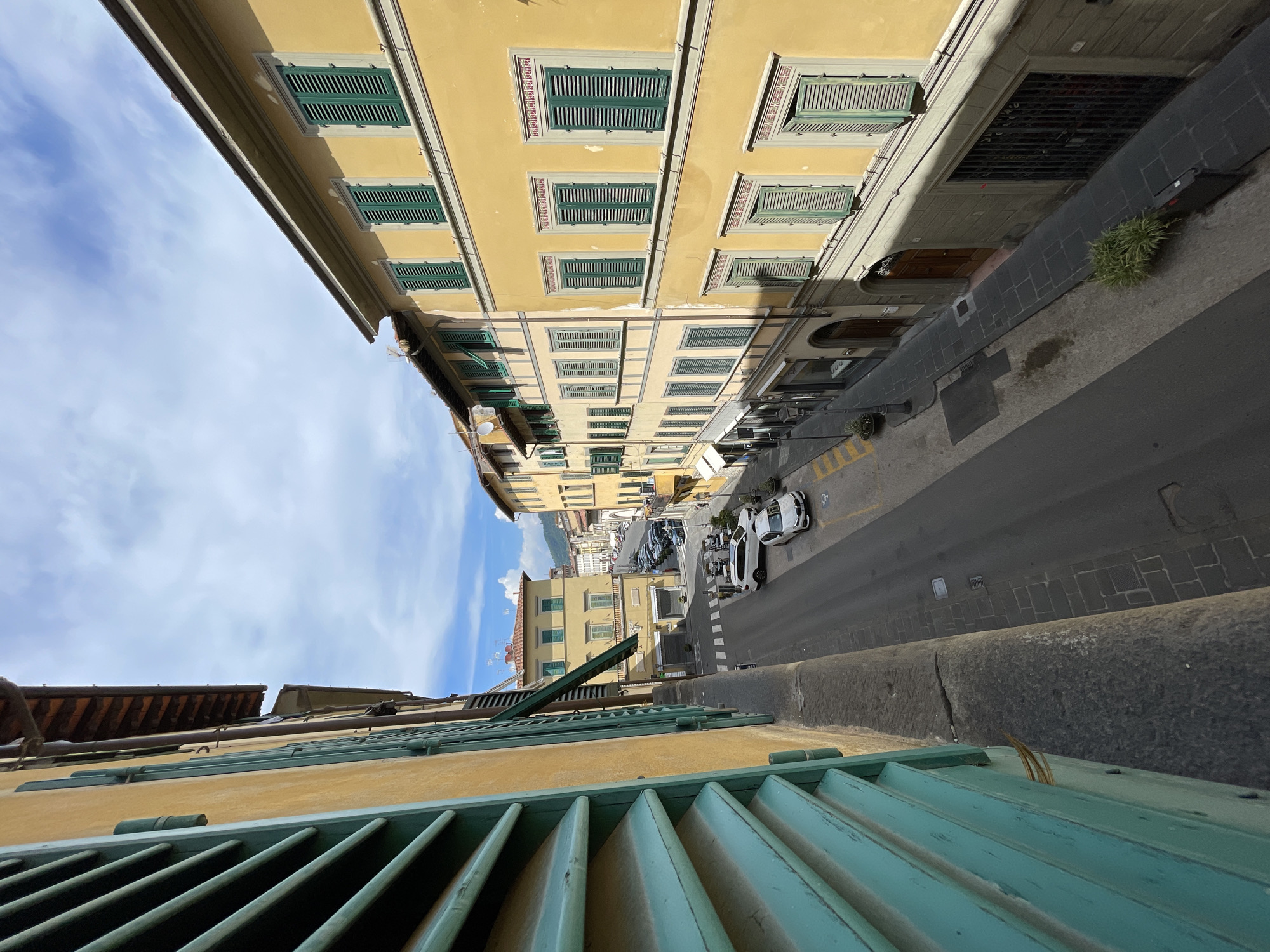 This is a high-angle, eye-level shot looking down a street in Pescia, Italy. The street is lined with buildings, mostly yellow, with green shutters. The sky is partly cloudy. In the foreground, to the left, is a large, partially open window with teal-colored shutters. The street is paved with asphalt and sidewalks are paved with stone. There are several cars parked along the street and one white car is driving down the street. The buildings appear to be residential or commercial, with shopfronts visible on the lower levels of some. There are some plants in pots along the sidewalks. The overall impression is of a quiet, sunny day in an Italian town.