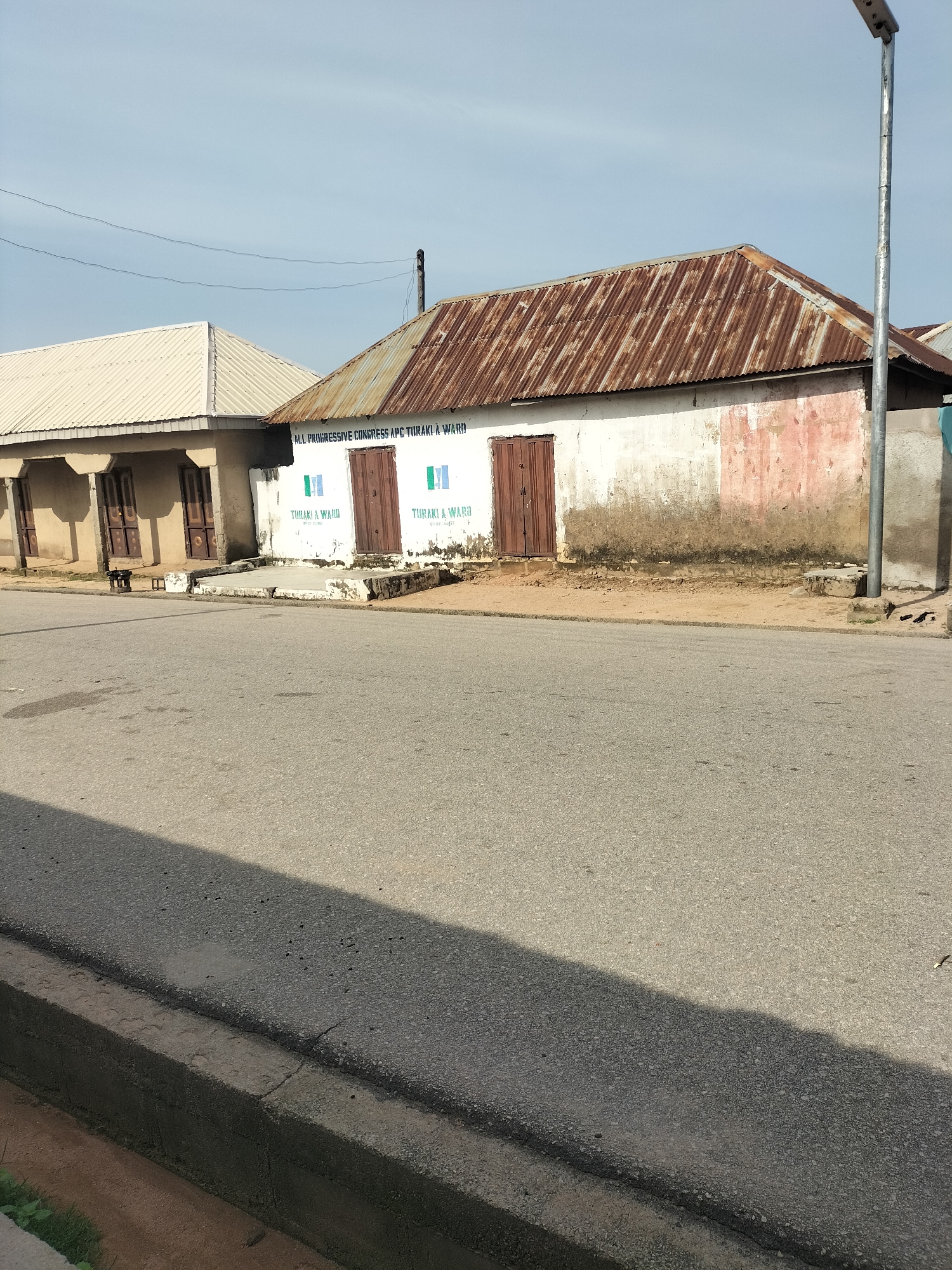A two-story building with a corrugated iron roof is visible on the right side of the image, in Jalingo, Nigeria. The building has white walls with some peeling pink paint and two brown wooden doors. Above the doors, the words "ALL PROGRESSIVE CONGRESS APC TURAKI & WARD" are written in green. Below the inscription, two Nigerian flags are painted. A street lamp is located to the right of the building. Another building with a lighter colored corrugated roof is visible to the left, with pillars and darker windows. The buildings are situated next to a paved road, with a curb and a patch of sandy dirt between the road and the buildings. The sky is clear and blue.
