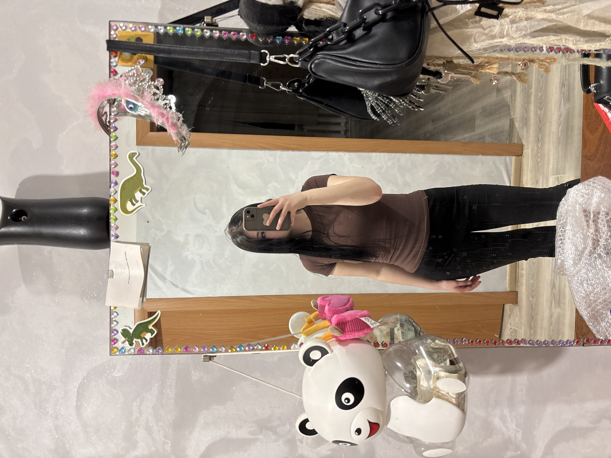 The image shows a reflection of a person in a mirror with a decorated frame. The person is holding a smartphone and appears to be taking a picture of themself. The person has long dark hair, and wears a brown t-shirt and black pants.
The mirror frame is decorated with colorful gemstones and stickers, including dinosaur figures. Hanging over the mirror are several items, including a black purse with a chain strap and other accessories. A panda-shaped object is attached near the bottom of the mirror, along with a transparent container filled with what appears to be money. The background is a wall with a textured surface, possibly a marble or stucco effect. The overall setting suggests an interior space, likely a private residence. There's no information regarding Фоки, Russia in this image.