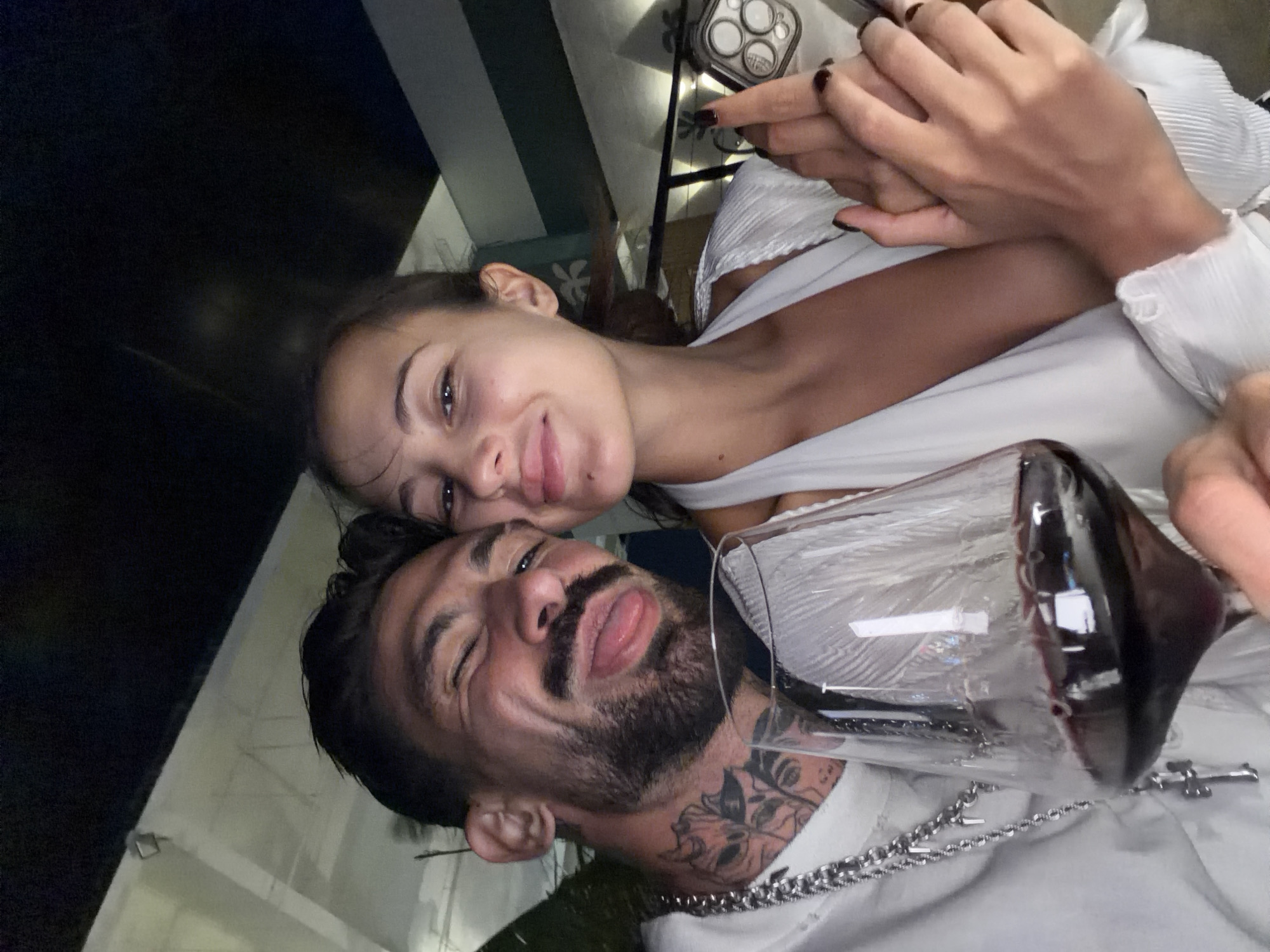A man and woman pose together in a selfie. The woman is fair-skinned with brown hair and is wearing a white top. The man has dark hair, a beard, and tattoos on his neck. He is wearing a white shirt and a silver chain. A large wine glass is in the foreground, partially obscuring the view of the man. The background appears to be an outdoor setting, possibly a patio or balcony. Based on the prompt, the location is Pattaya City, Thailand.
