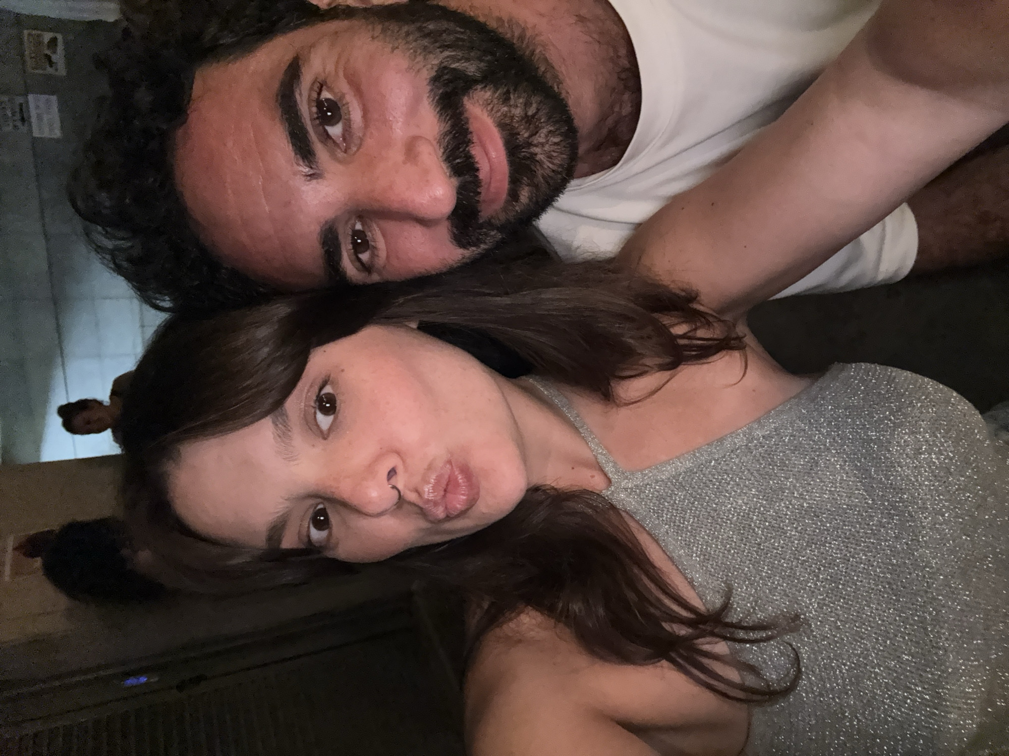 This is a selfie taken by a couple in Tel Aviv, Israel. The woman is in the foreground, her face close to the camera, with her lips puckered as if for a kiss. She has dark hair and freckles. She is wearing a sparkly silver halter top. Beside her, the man is looking at the camera with a slight smile. He has dark, curly hair and a beard. He is wearing a white t-shirt. In the background, blurred figures can be seen, suggesting they are in a public place, possibly a street or a venue with other people present. The lighting is dim, indicative of an evening or indoor setting.