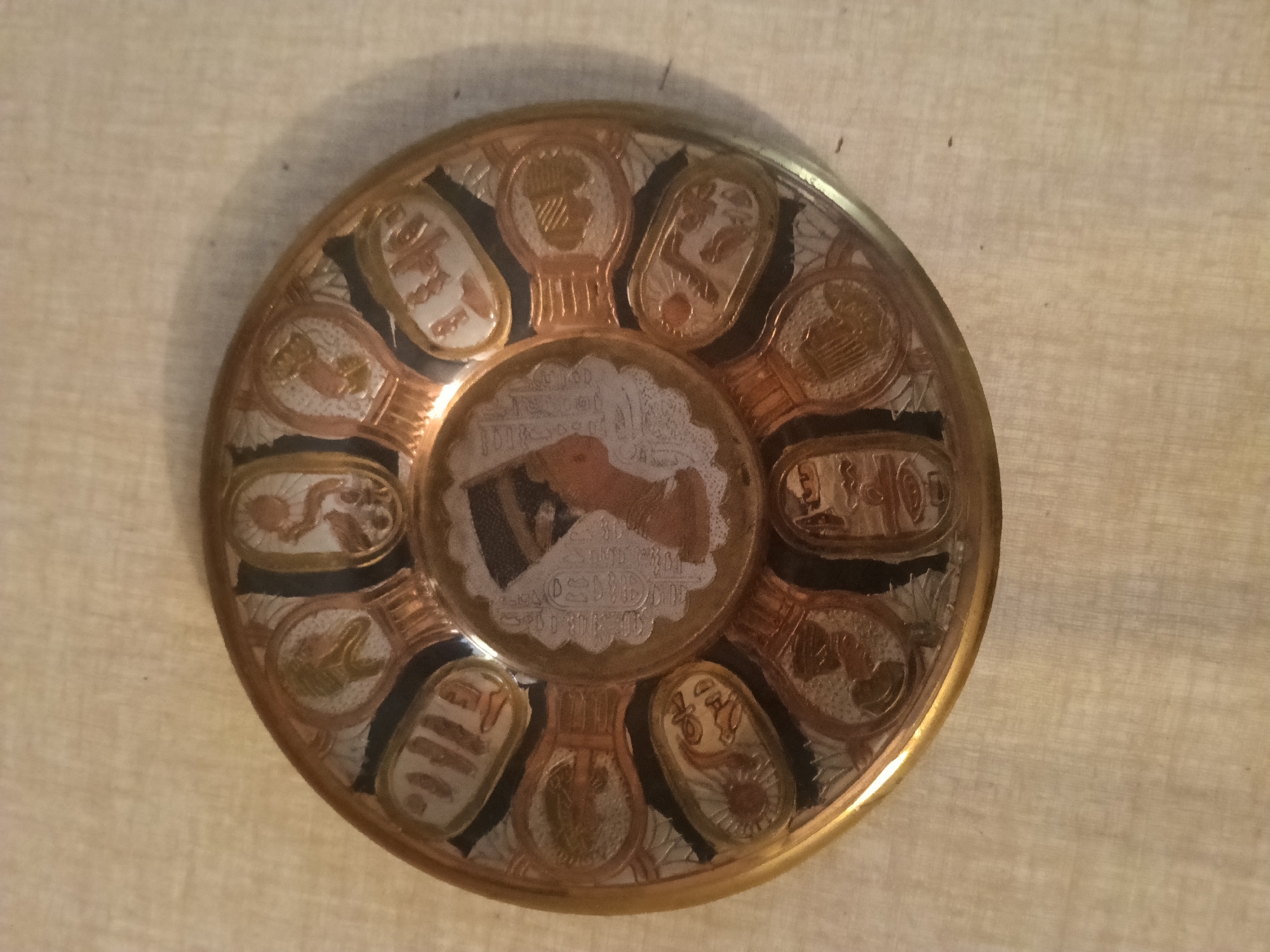A circular metal plate with Egyptian motifs is centered on a textured, light-colored surface, possibly fabric. The plate features a golden rim and is decorated with various designs in gold, silver, and black. The central part of the plate depicts a scene with figures and hieroglyphs. Around the center, there are multiple oval-shaped cartouches, each containing different symbols or figures. These cartouches are arranged in a radial pattern. The overall style suggests an artifact or souvenir with Egyptian-inspired artwork. The background is uniformly lit, casting a slight shadow to the left of the plate. The image does not contain any identifiable location in Rybnitsa, Moldova.