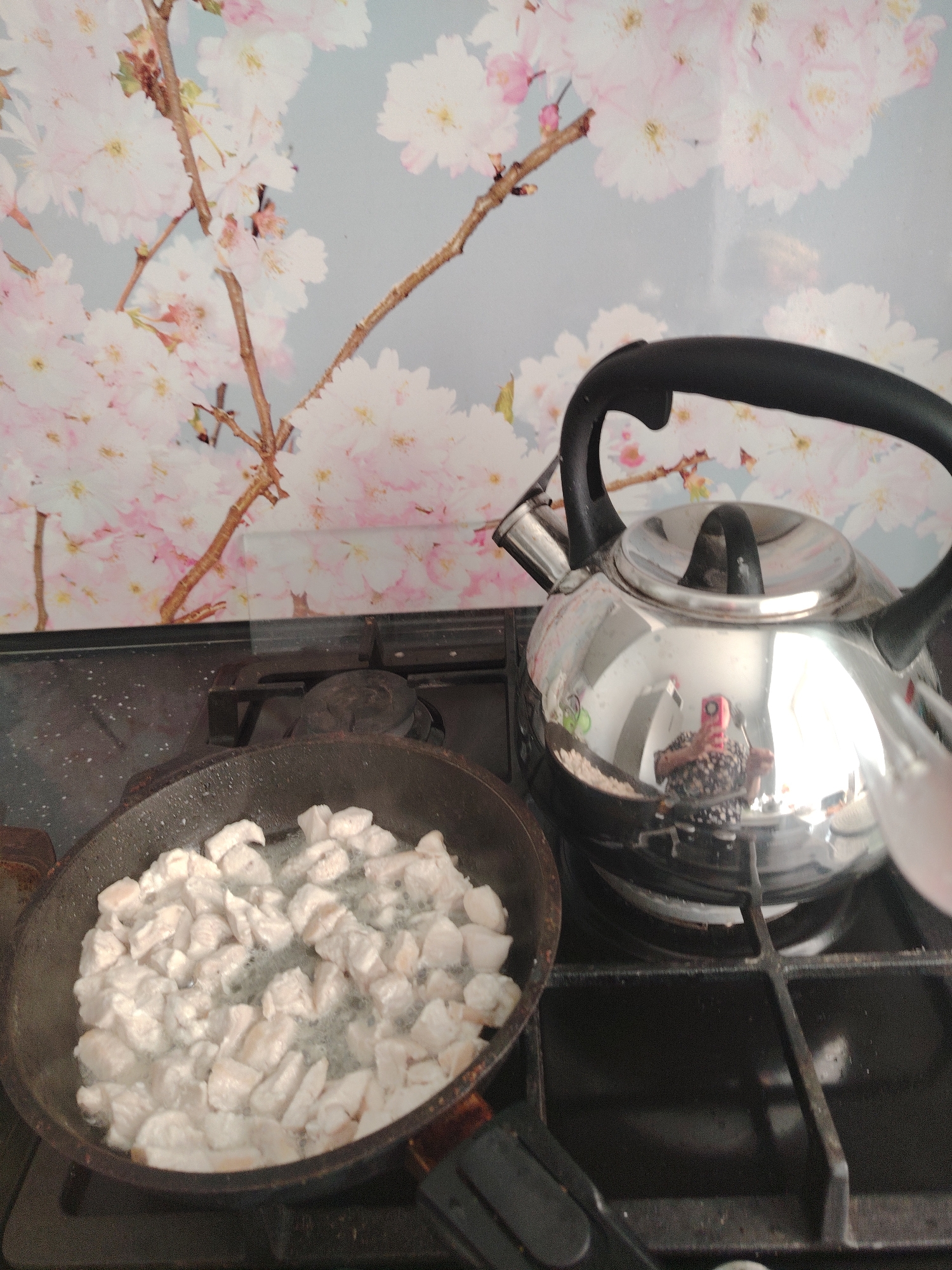 A top-down view of a kitchen scene in what appears to be Sarapul, Russia, is captured. The scene features a black gas stovetop. A dark, well-used frying pan on the left contains small, white cubes of food being cooked, likely chicken. To the right of the pan sits a silver, reflective kettle with a black handle. The backdrop consists of a floral patterned splashback, resembling pink cherry blossoms. The kettle reflects the person taking the picture. The objects are arranged on the stovetop, which appears slightly worn.
