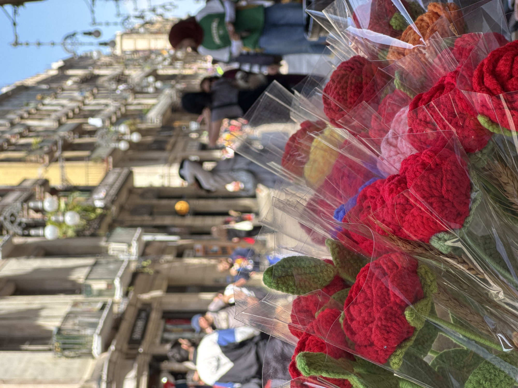 The image captures a vibrant street scene, with a primary focus on colorful crocheted flowers in the foreground. The photograph is oriented vertically, showing a close-up of several bouquets of artificial flowers, mainly bright red roses with green leaves. Each bouquet is individually wrapped in clear plastic and appears to include stalks of dried wheat. Other colors like yellow, pink, orange, and blue are visible in additional bouquets, suggesting a variety of options.
In the blurred background, a bustling street or plaza in Barcelona, Spain, unfolds. Tall, multi-story buildings with balconies line the thoroughfare, hinting at classic European architecture. Numerous people are visible walking along the street, though their faces and specific activities are indistinct due to the shallow depth of field. One person in the midground wears a green vest over a white shirt and blue jeans, topped with a reddish-brown hat. Street decorations, possibly lights or banners, are strung across the street overhead, with one distinctive golden orb-like decoration hanging prominently.
The scene takes place during the daytime under a bright, clear sky, indicating good weather. The overall atmosphere appears lively and cheerful, likely a commercial or tourist area where these handmade flowers are being sold. There is no legible text visible within the image.