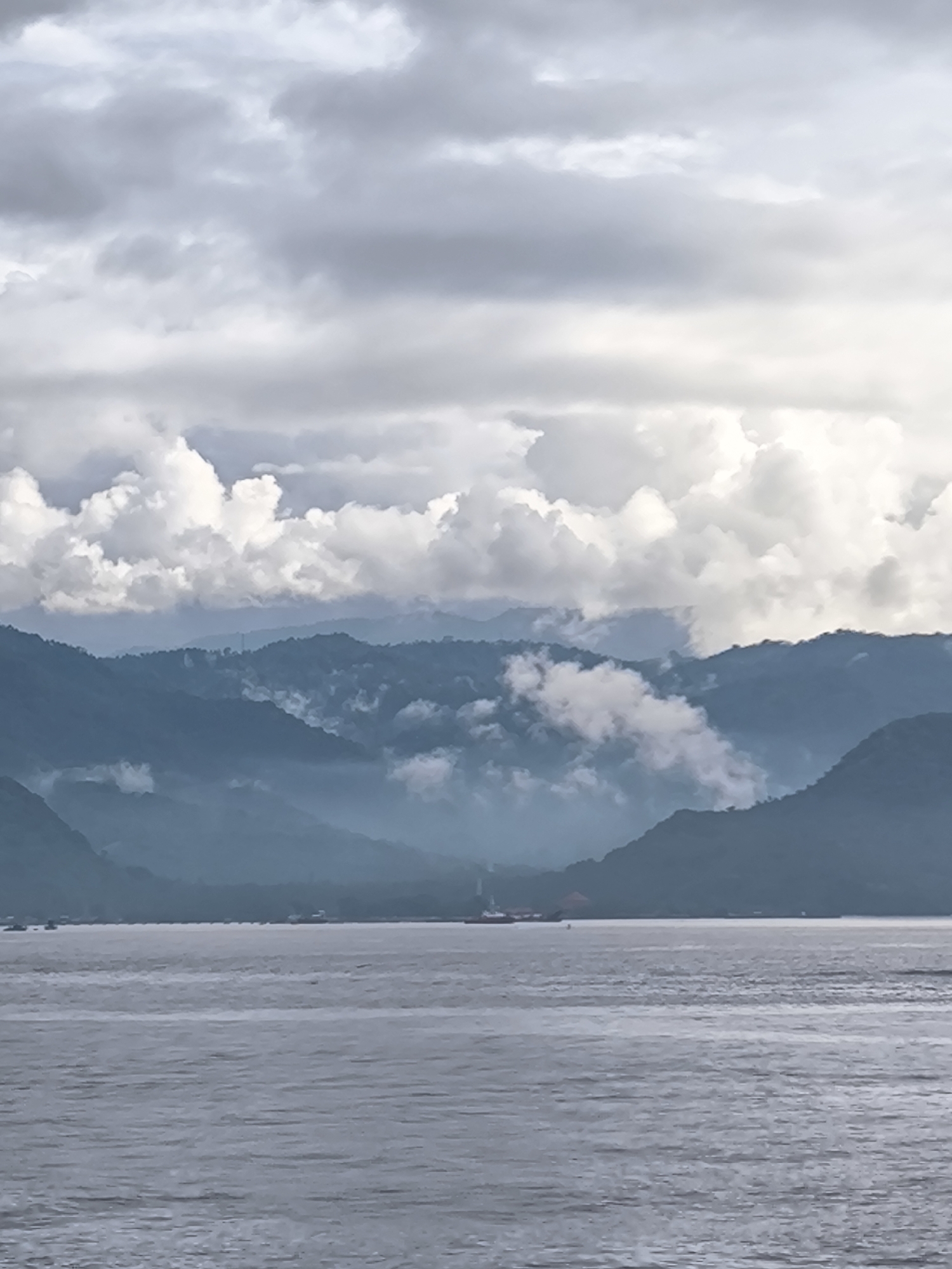 The image presents a vast and tranquil natural landscape, primarily composed of water, mountains, and a cloudy sky. In the foreground, a wide expanse of calm, dark grey water occupies the lower third of the frame, showing subtle ripples and reflections.
Above the water, the middle ground is dominated by multiple layers of lush, dark green mountains and hills. These mountains recede into the distance, with their lower slopes and valleys partially obscured by thick, white and grey mist or low-hanging clouds. This atmospheric haze creates a sense of depth and mystery, with some mountain peaks intermittently appearing through the swirling clouds. Along the distant waterline, near the base of the mountains, several small, indistinct vessels are visible. A slightly larger, reddish-orange boat or structure can be faintly discerned just above the horizon line, slightly left of the center.
The upper portion of the image is filled with an overcast sky. Heavy clouds of varying shades, from bright white and fluffy near the mountain tops to darker, more uniform grey higher up, dominate the celestial expanse. The diffused lighting suggests a cloudy day, making the exact time of day ambiguous, though it is clearly daylight.
The scene is devoid of overt human activity, conveying a sense of serene natural beauty and vastness. The prevailing weather conditions include heavy cloud cover and mist blanketing the mountainous terrain. There is no visible text in the image. The layered topography and coastal appearance are consistent with island or coastal regions, fitting the provided context of Indonesia.