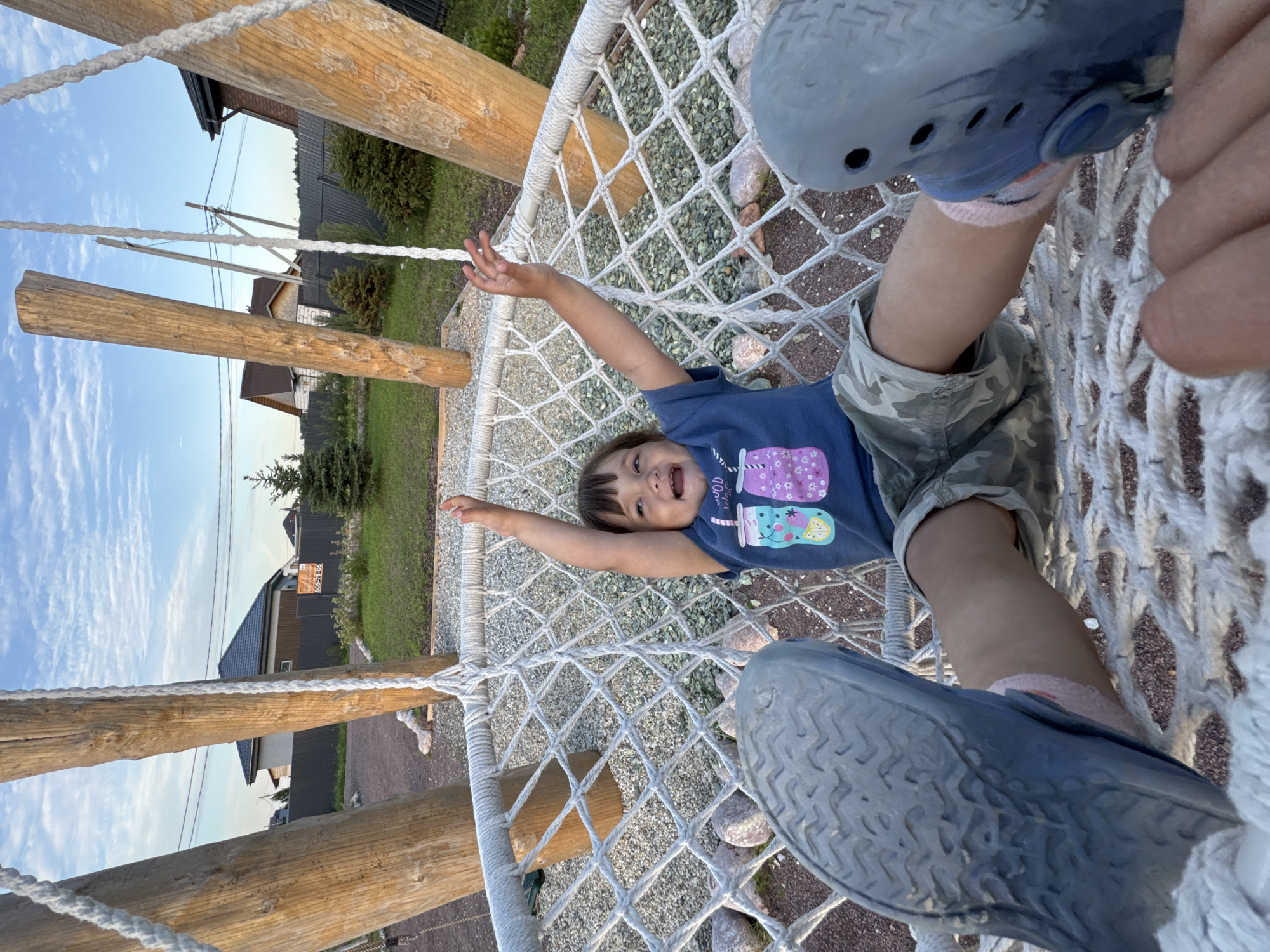 A young child lies on a net swing, looking up at the camera with a wide smile and arms outstretched. The child is wearing a blue t-shirt with a graphic of drinks and camouflage shorts. Their feet are clad in blue shoes, and they are wearing pink socks. The swing is suspended from a wooden frame. In the background, houses and trees are visible under a partly cloudy sky, suggesting it is daytime. The overall mood is cheerful and playful. The image appears to have been taken in Kempelevo, Russia, as indicated by the context provided.